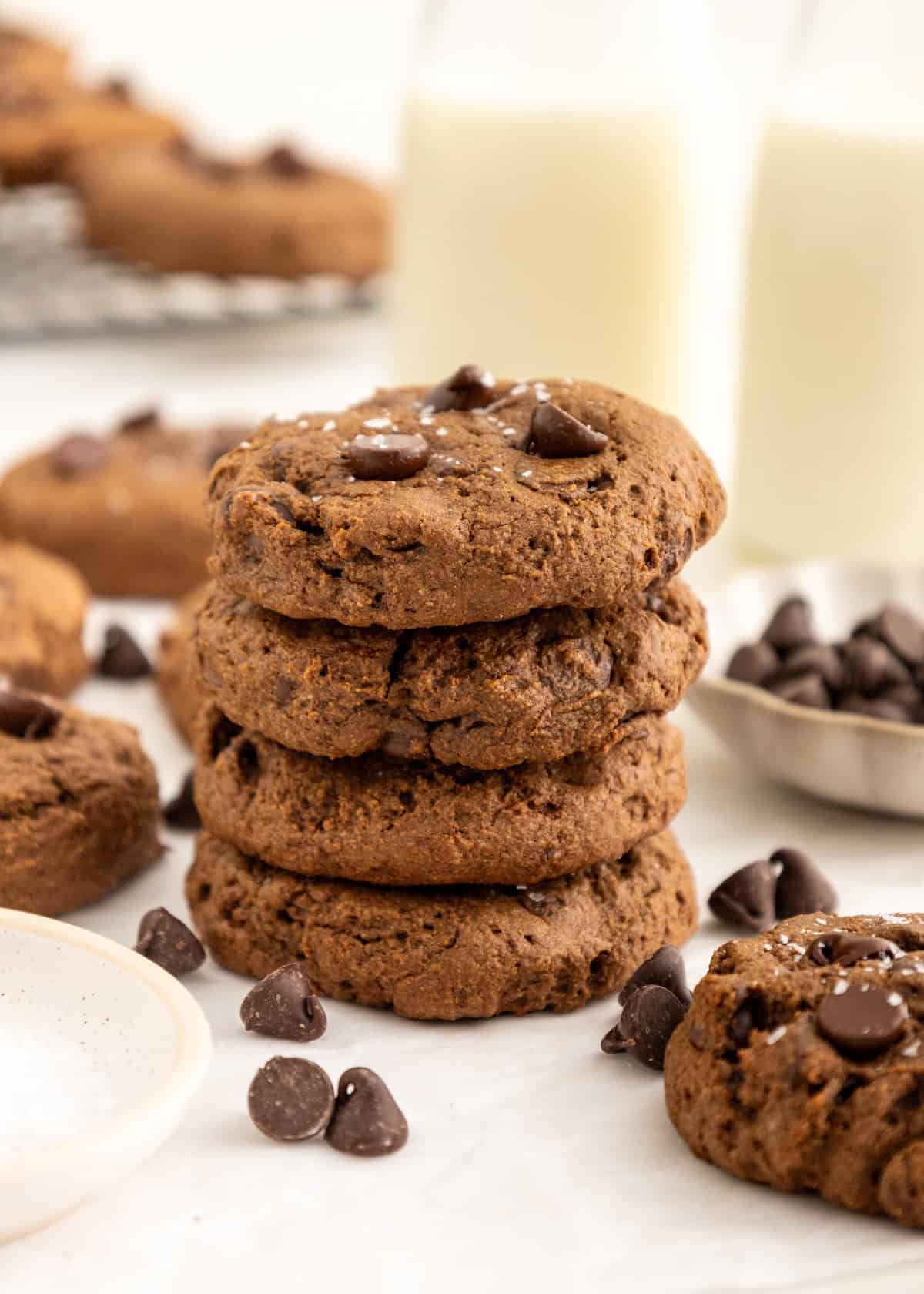 Stack of chocolate chip cookies with scattered chips and bottles of milk in the background.