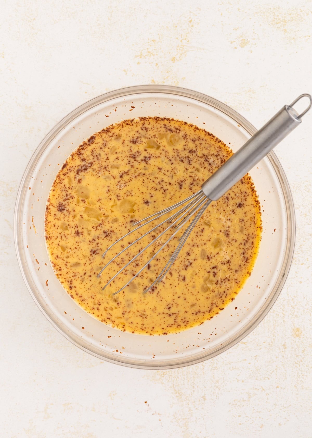 Glass bowl with yellow batter and a whisk, sprinkled with cinnamon, on a light surface.