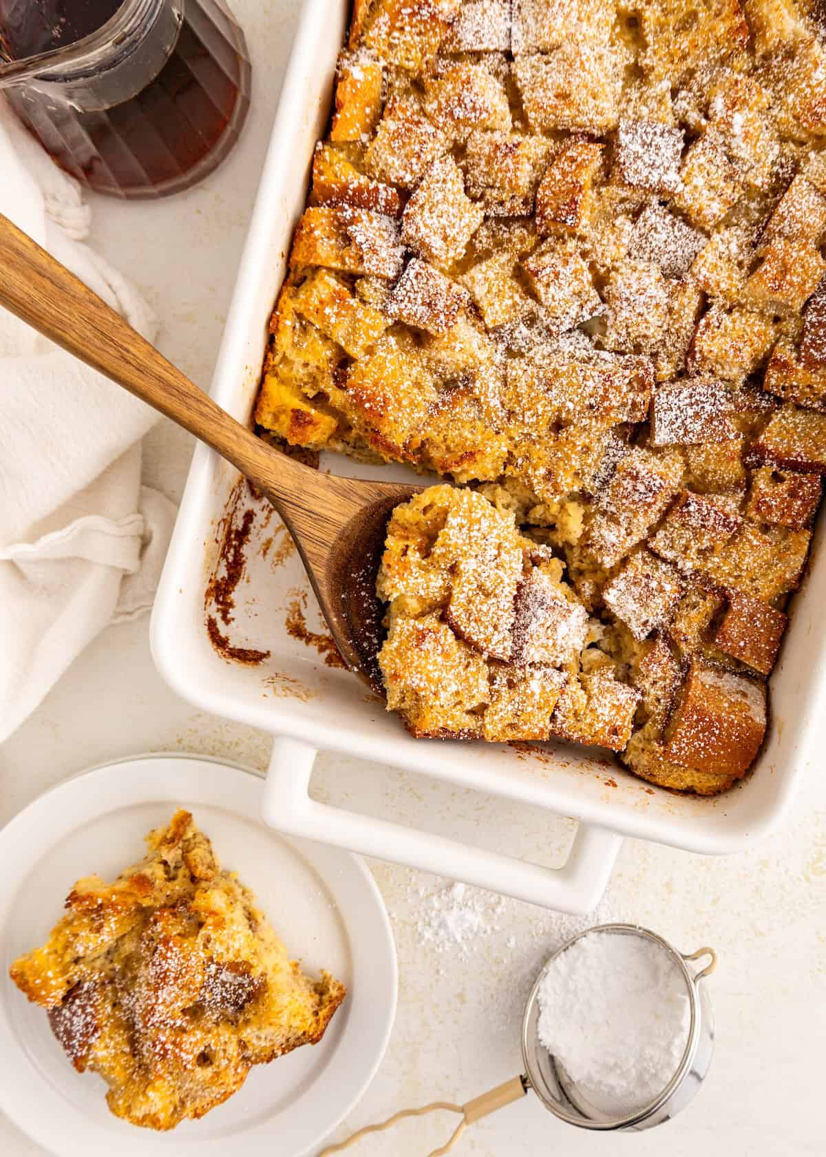 Bread pudding in a white baking dish, served on a plate, topped with powdered sugar.