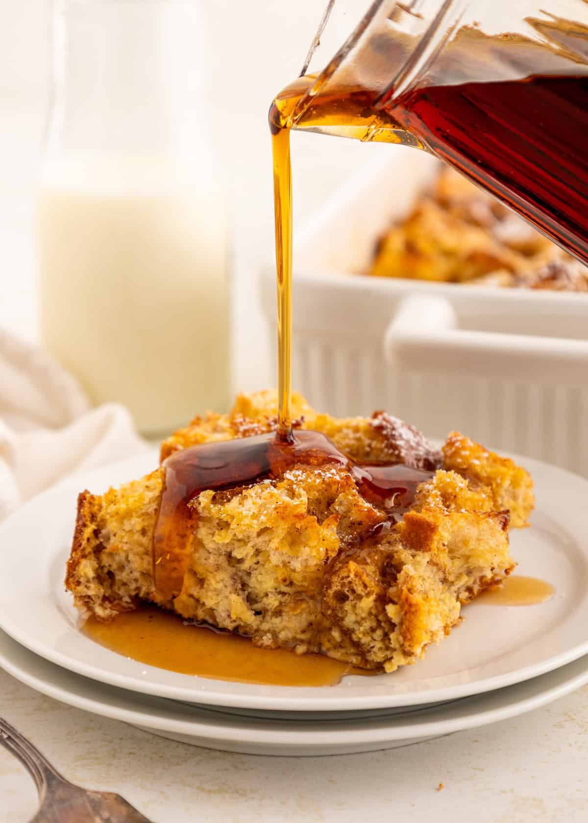 Syrup being poured over a slice of baked French toast on a white plate.