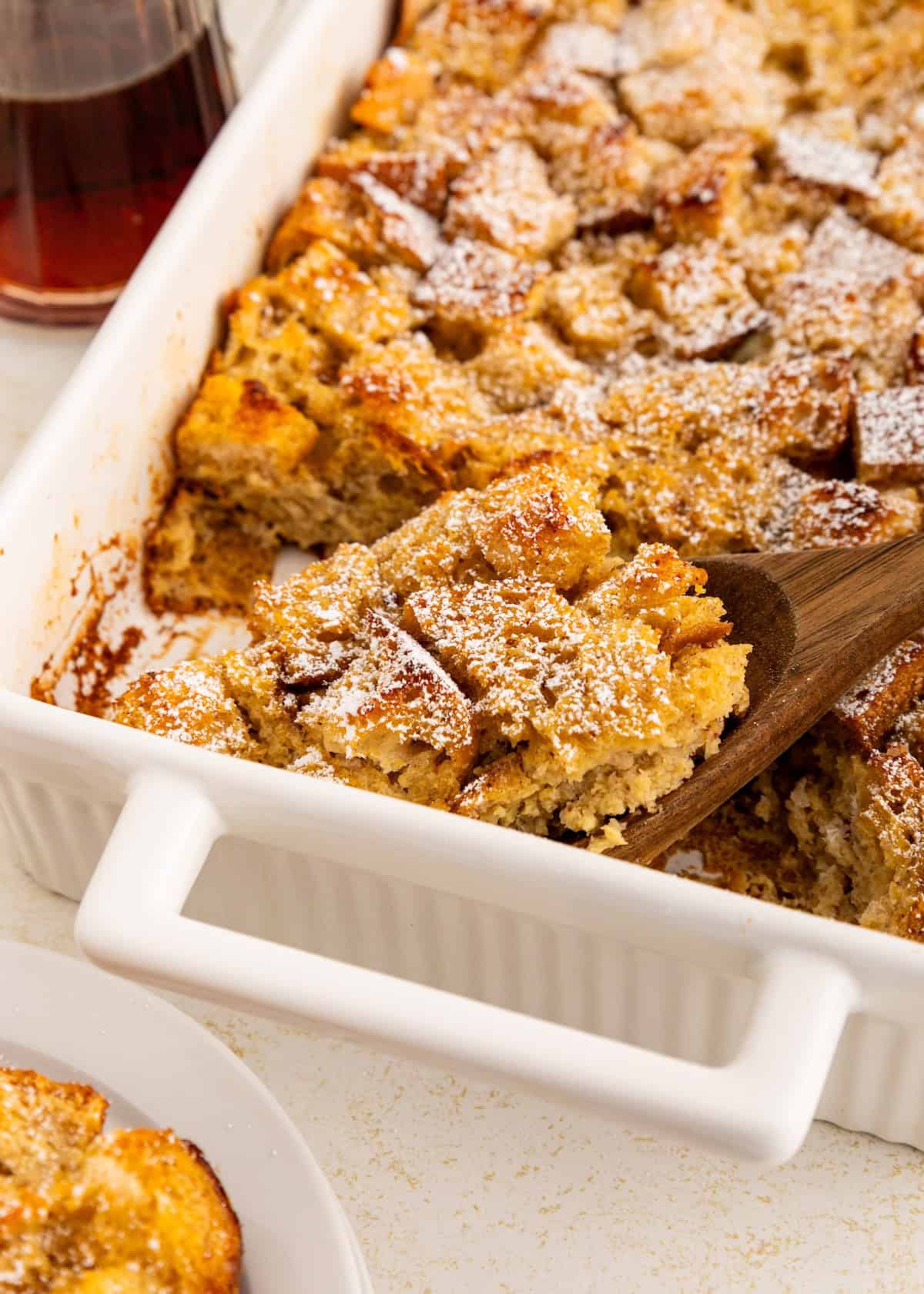 A wooden spoon serves baked French toast dusted with powdered sugar from a white casserole dish.