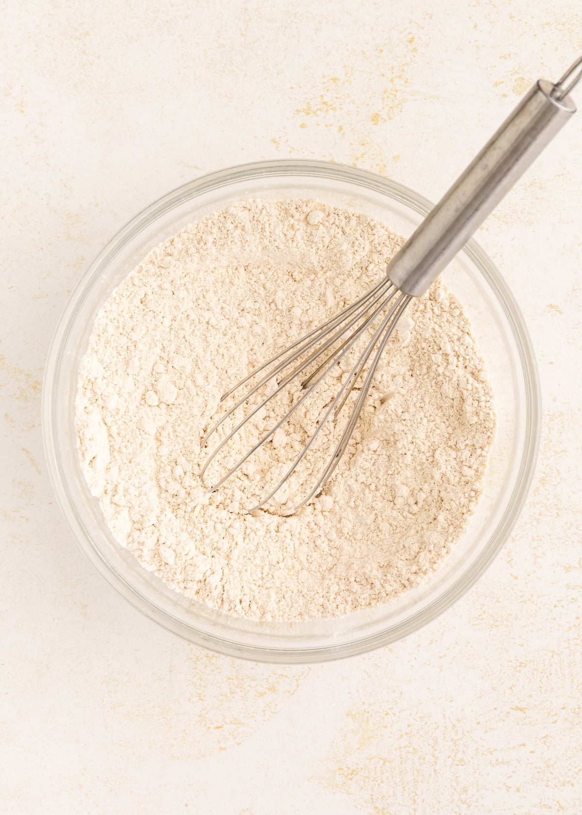 A metal whisk in a glass bowl filled with flour on a light surface.