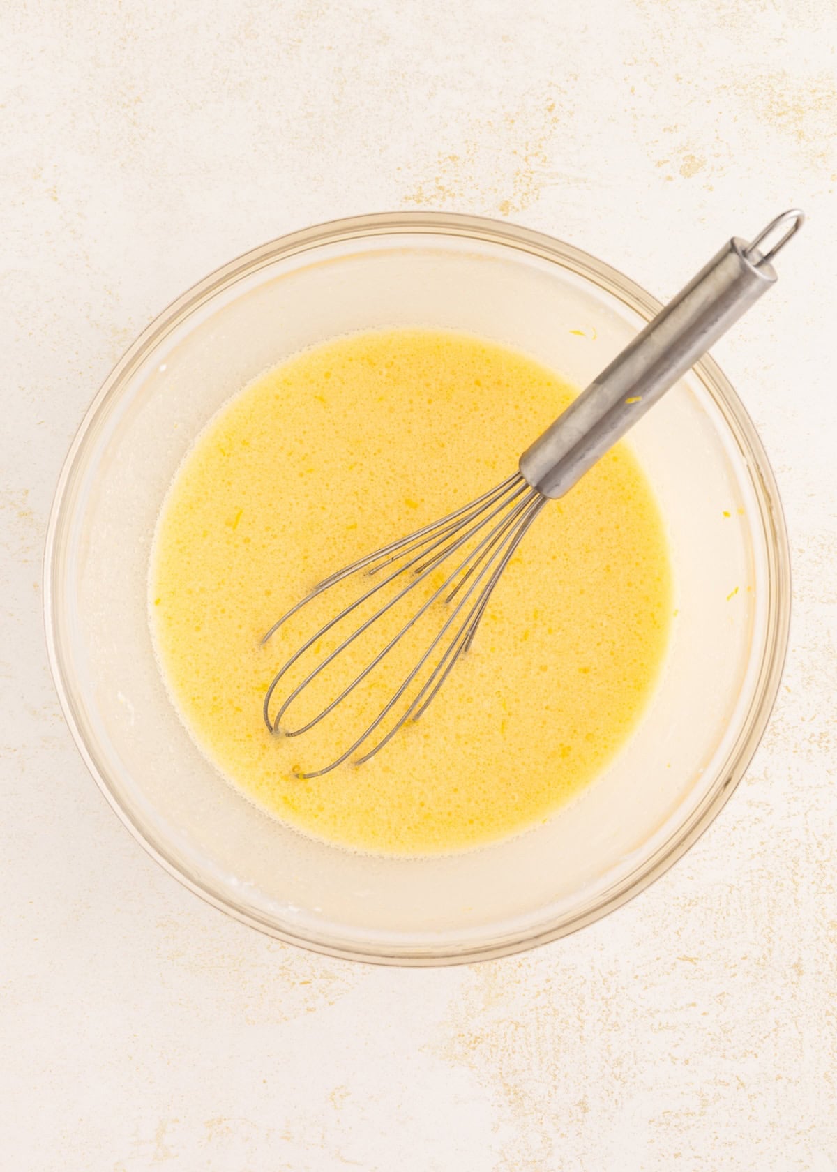 A whisk in a bowl of yellow batter on a light surface.