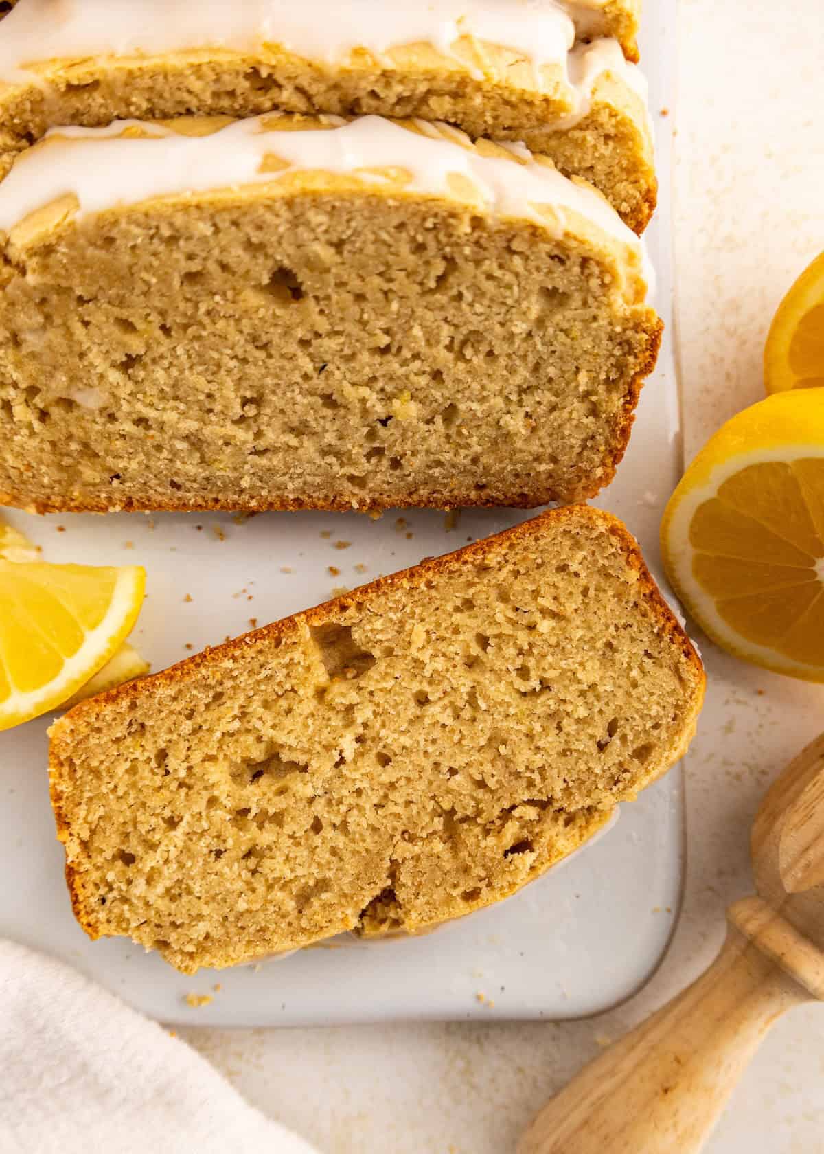 Sliced lemon loaf cake with glaze next to lemon wedges on a white surface.