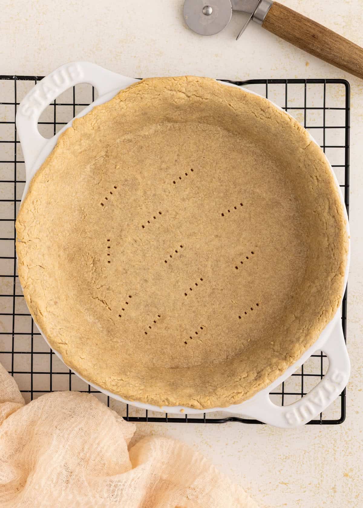 Unbaked pie crust in a white dish on a cooling rack, with a pastry cutter nearby.
