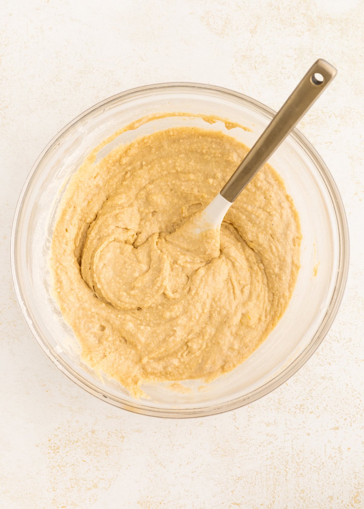 A glass bowl with thick, beige batter and a metal spoon on a light surface.