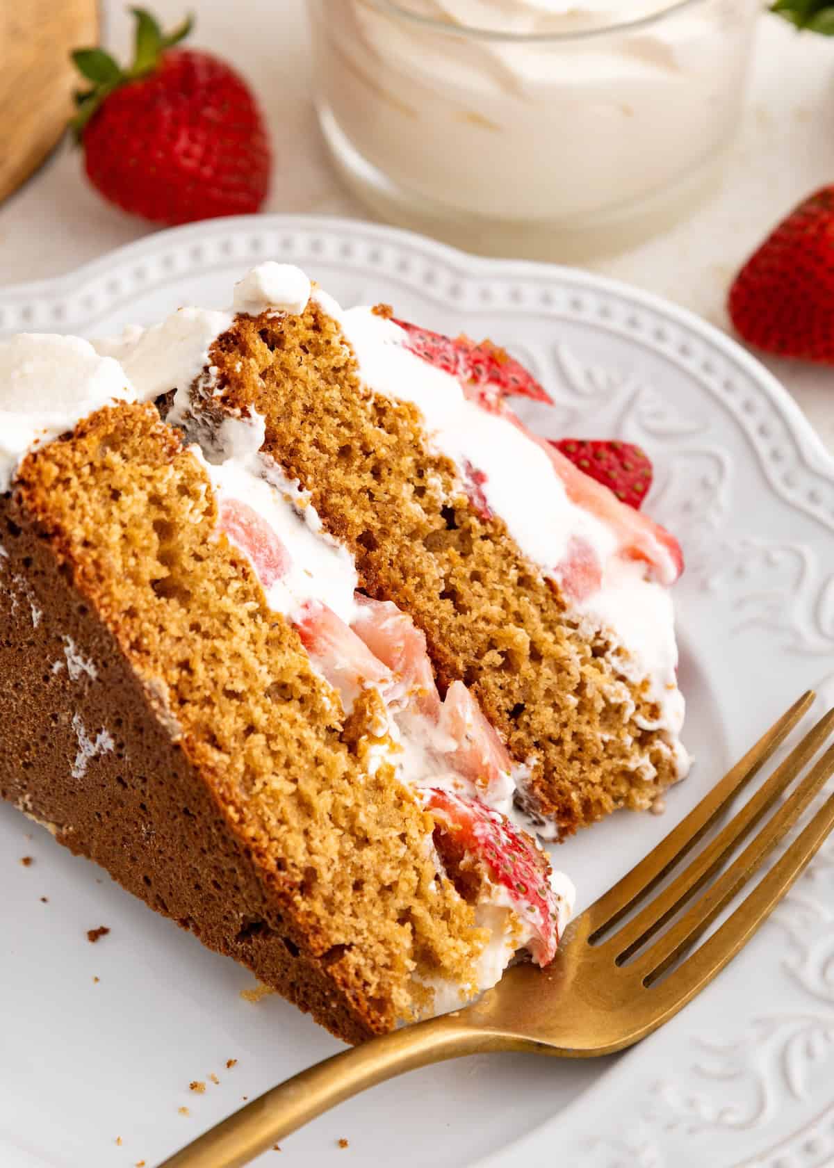 A slice of strawberry shortcake with whipped cream on a white plate, next to a gold fork.