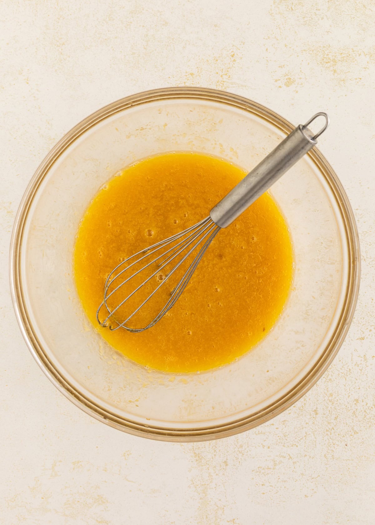 A metal whisk in a glass bowl with a yellow liquid mixture on a light surface.
