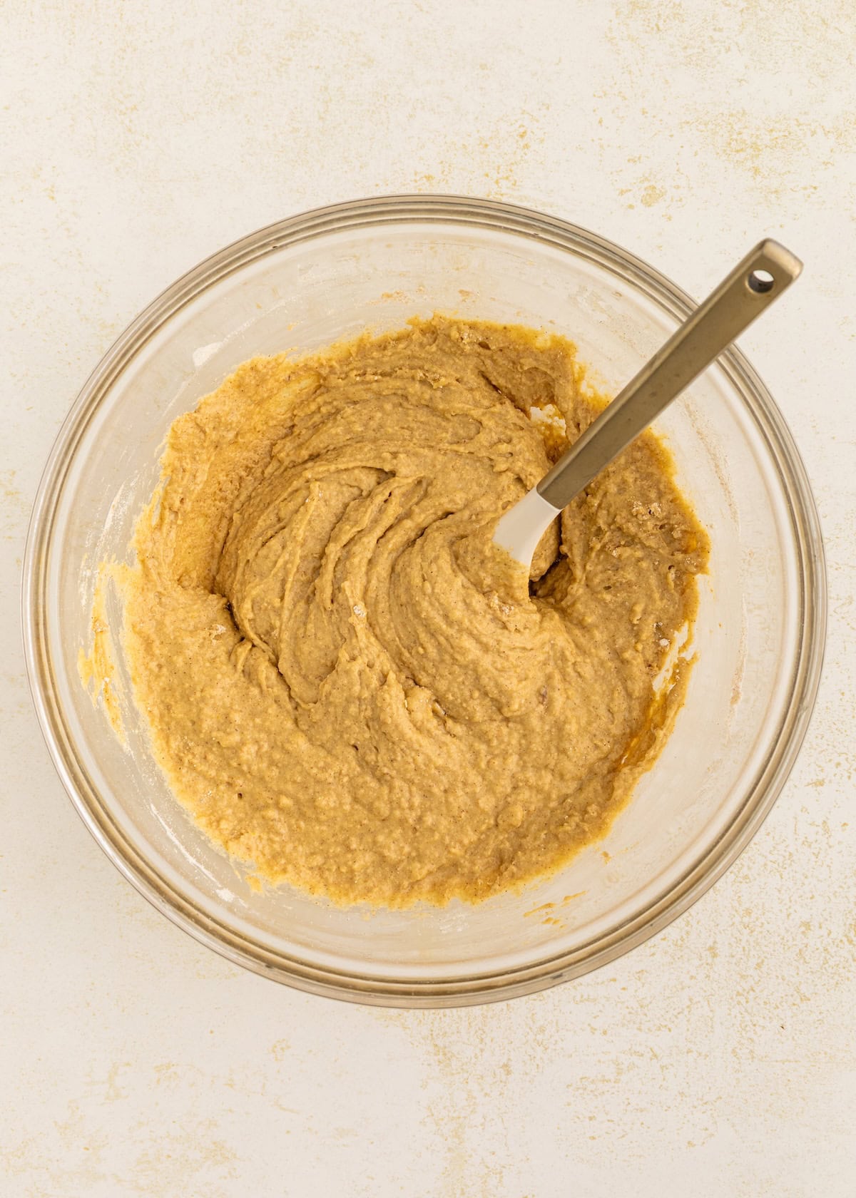 A glass bowl of light brown batter with a spatula on a light surface.