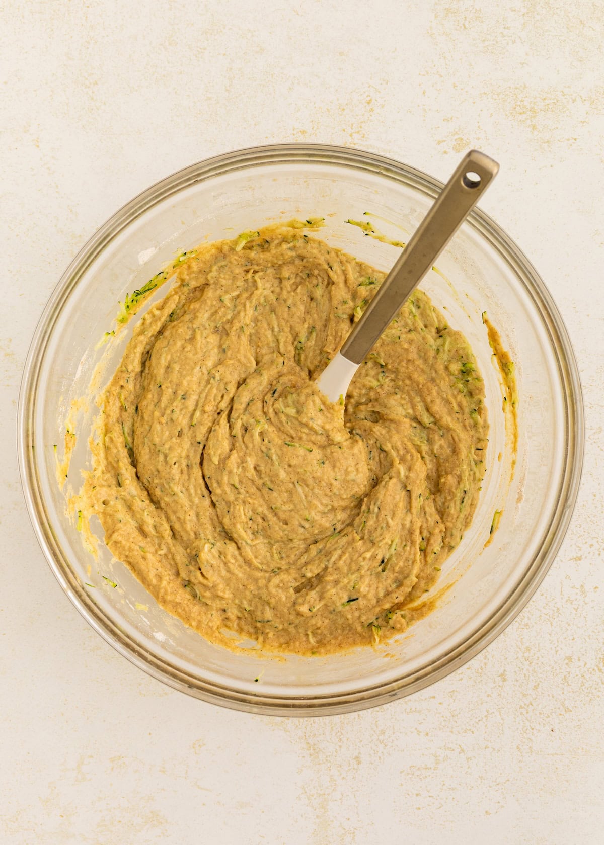 A glass bowl of zucchini bread batter with a spatula on a light surface.
