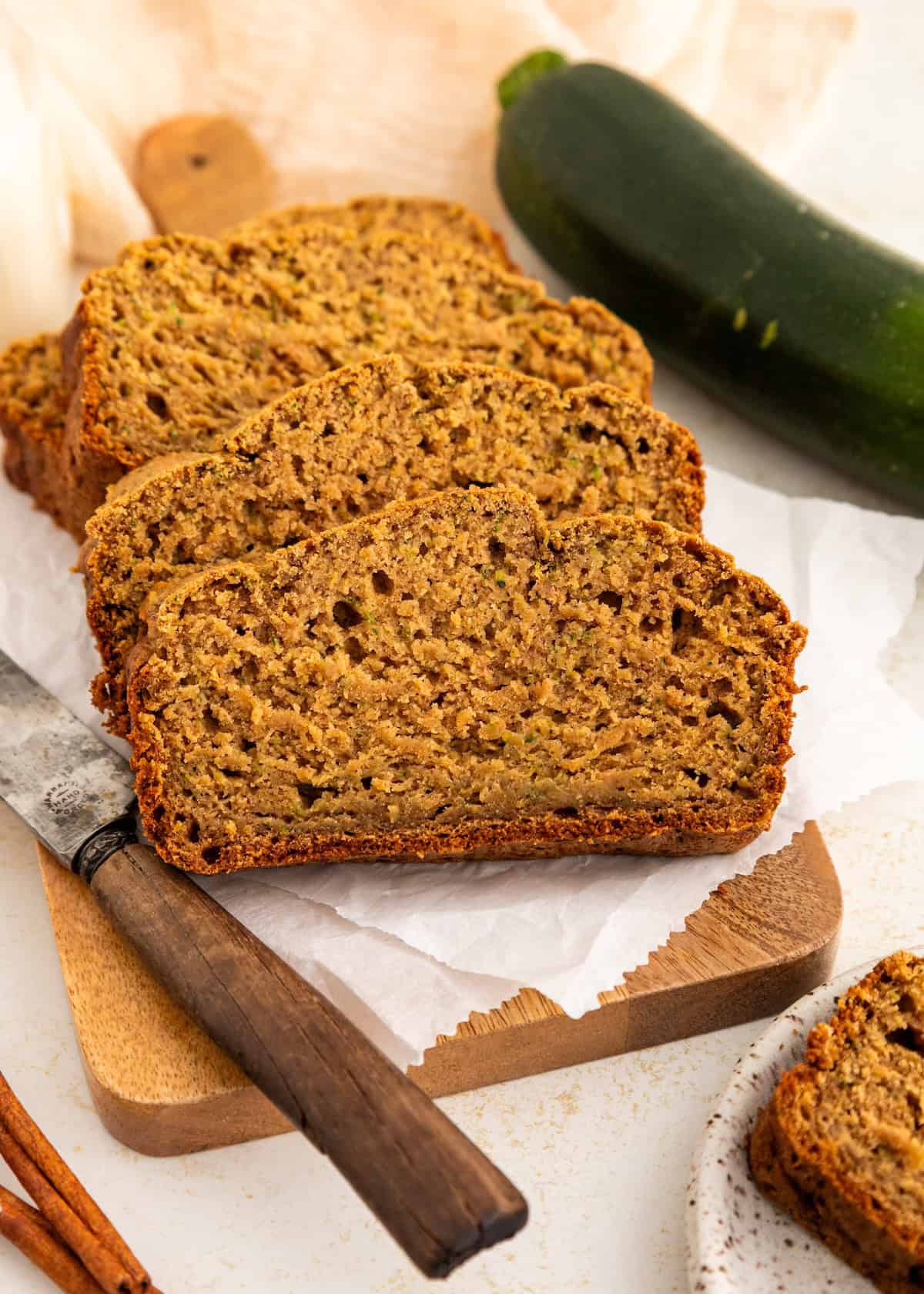 Three slices of zucchini bread on parchment paper with a knife and a whole zucchini nearby.