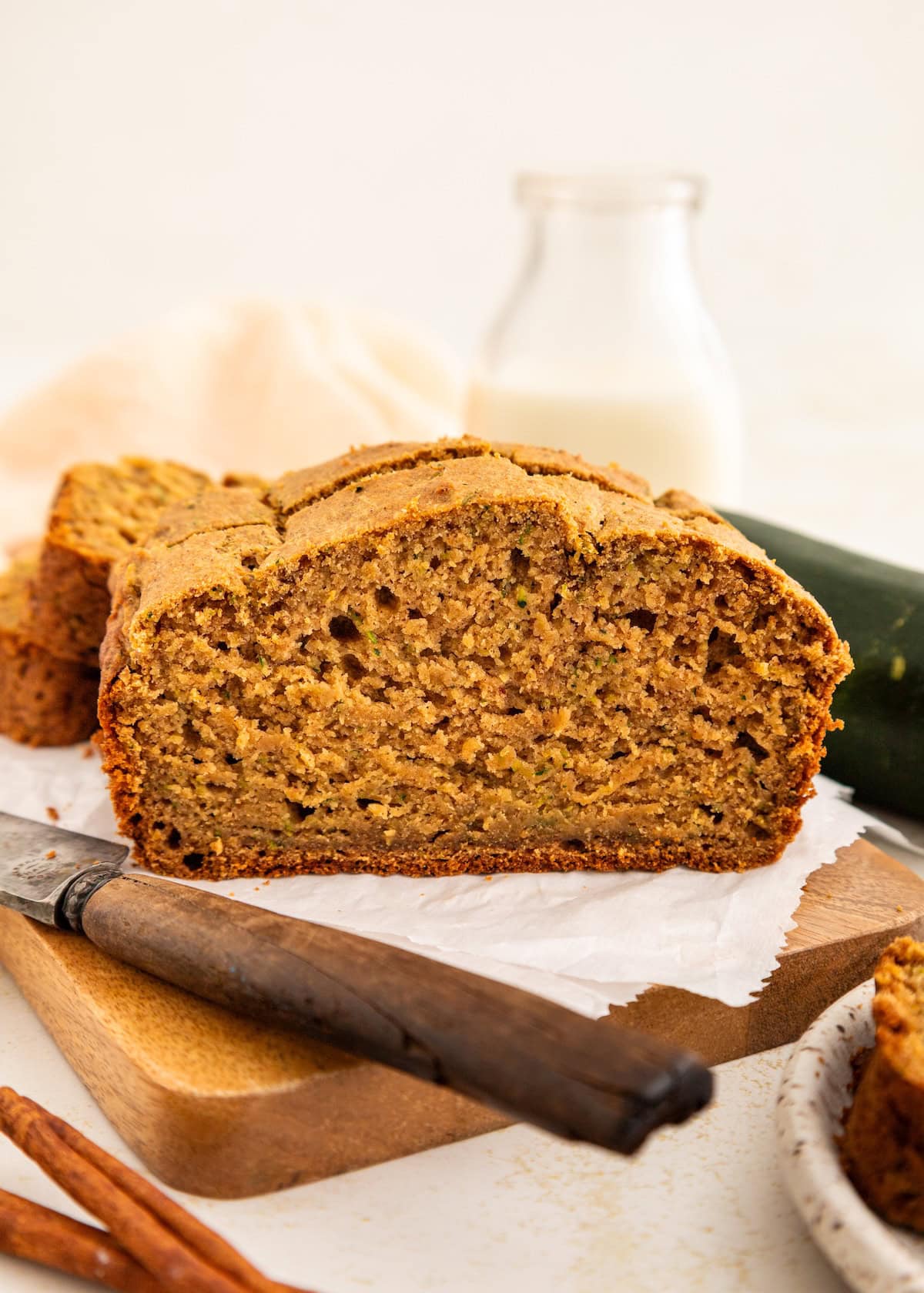 A sliced loaf of zucchini bread on parchment paper with a knife and a bottle of milk.