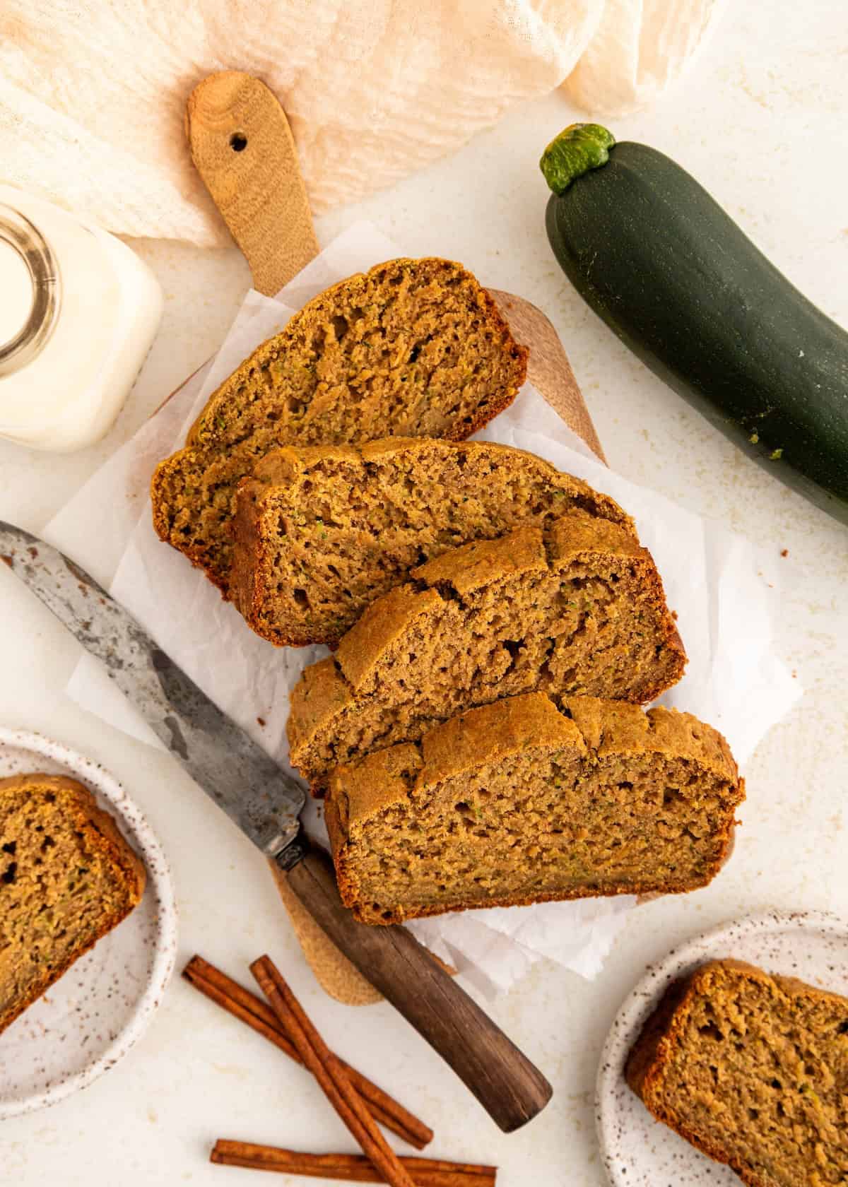 Sliced zucchini bread on a cutting board with a knife, zucchini, and cinnamon sticks nearby.