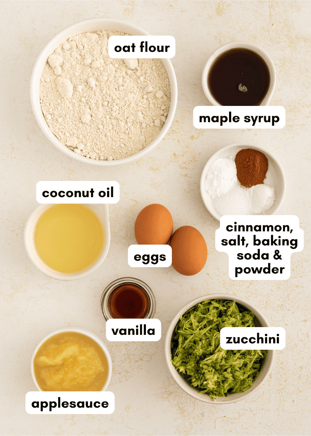 Labeled baking ingredients in bowls, including oat flour, eggs, zucchini, and others on a counter.