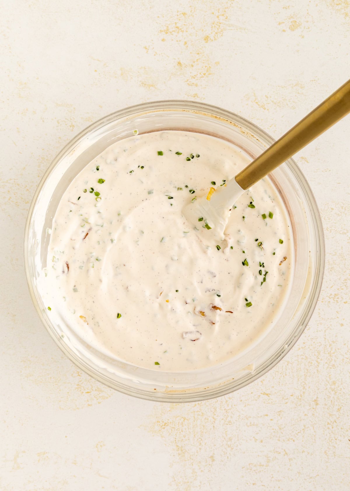 Bowl of creamy white dip with herbs, stirred by a gold-colored spoon.