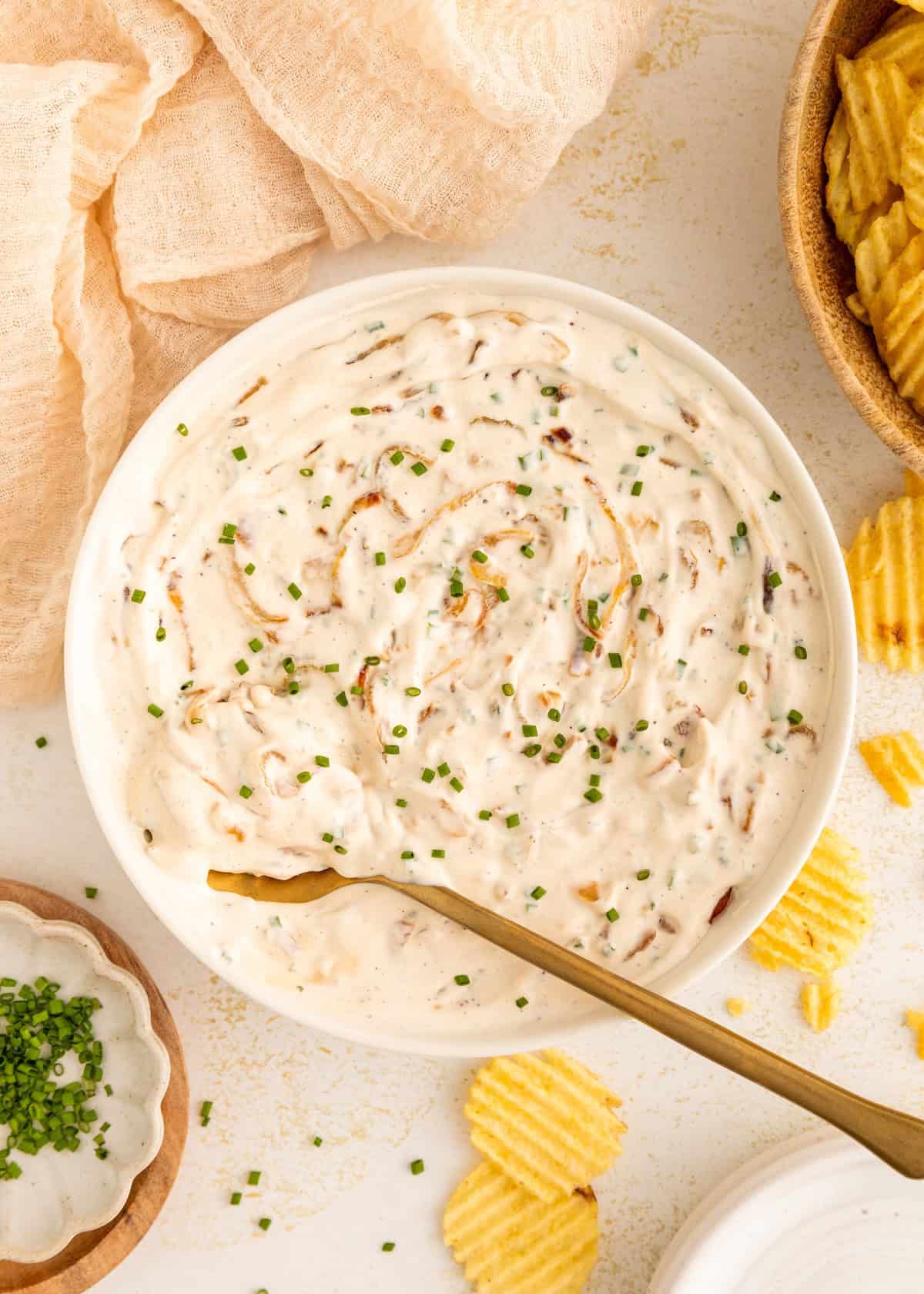 A bowl of creamy onion dip with chopped chives, surrounded by ridged potato chips.