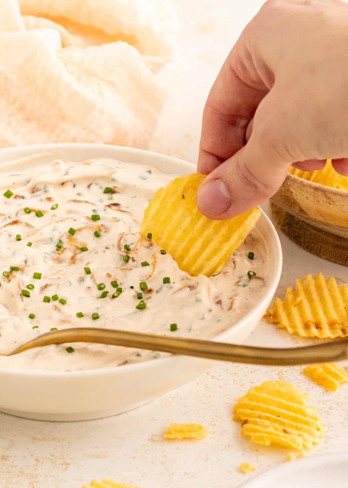 Hand dipping a ridged potato chip into creamy dip with chives in a white bowl.