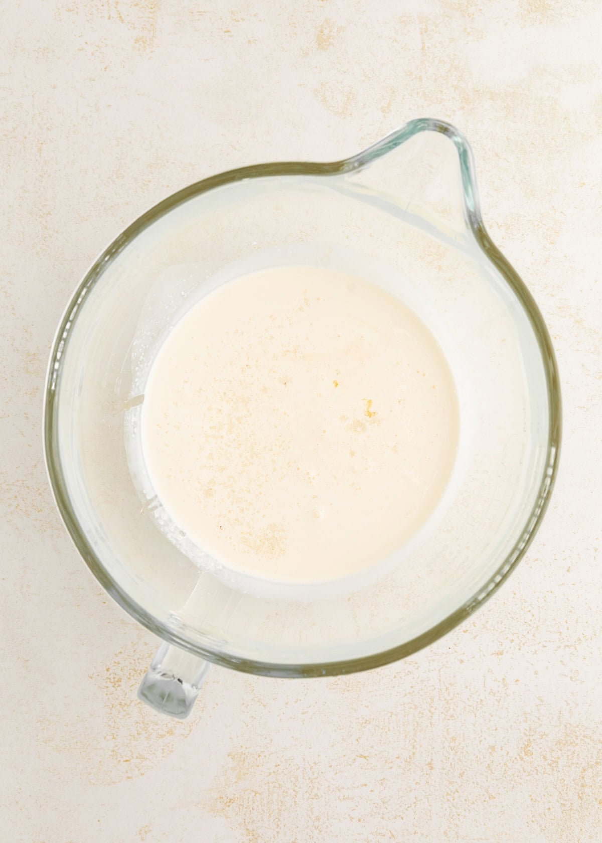 Glass mixing bowl with cream-colored liquid on a light, textured surface.