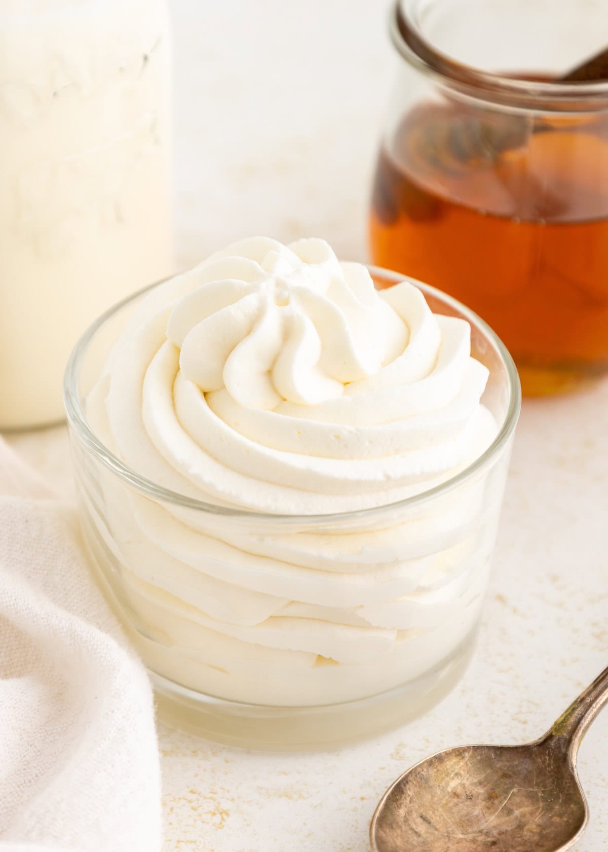 A glass bowl filled with swirled whipped cream, next to a spoon and a jar of honey.