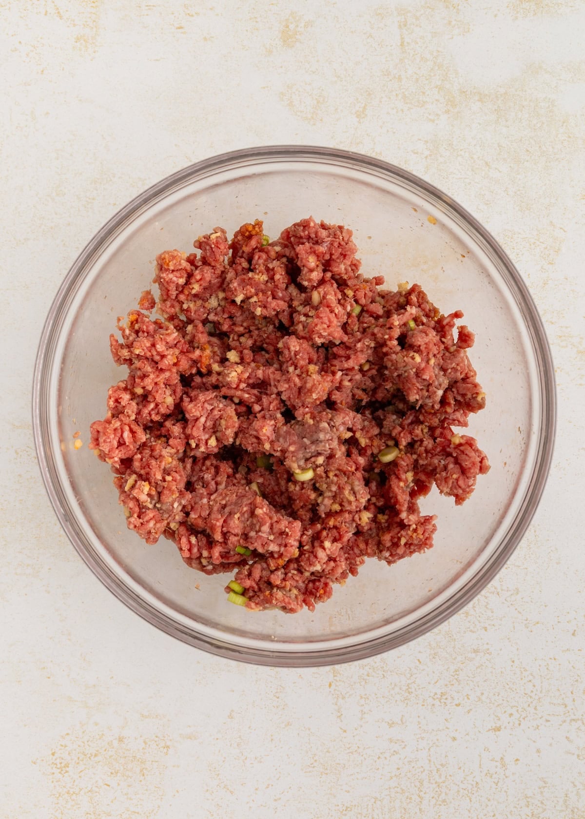 A glass bowl filled with seasoned raw ground beef mixture on a light surface.