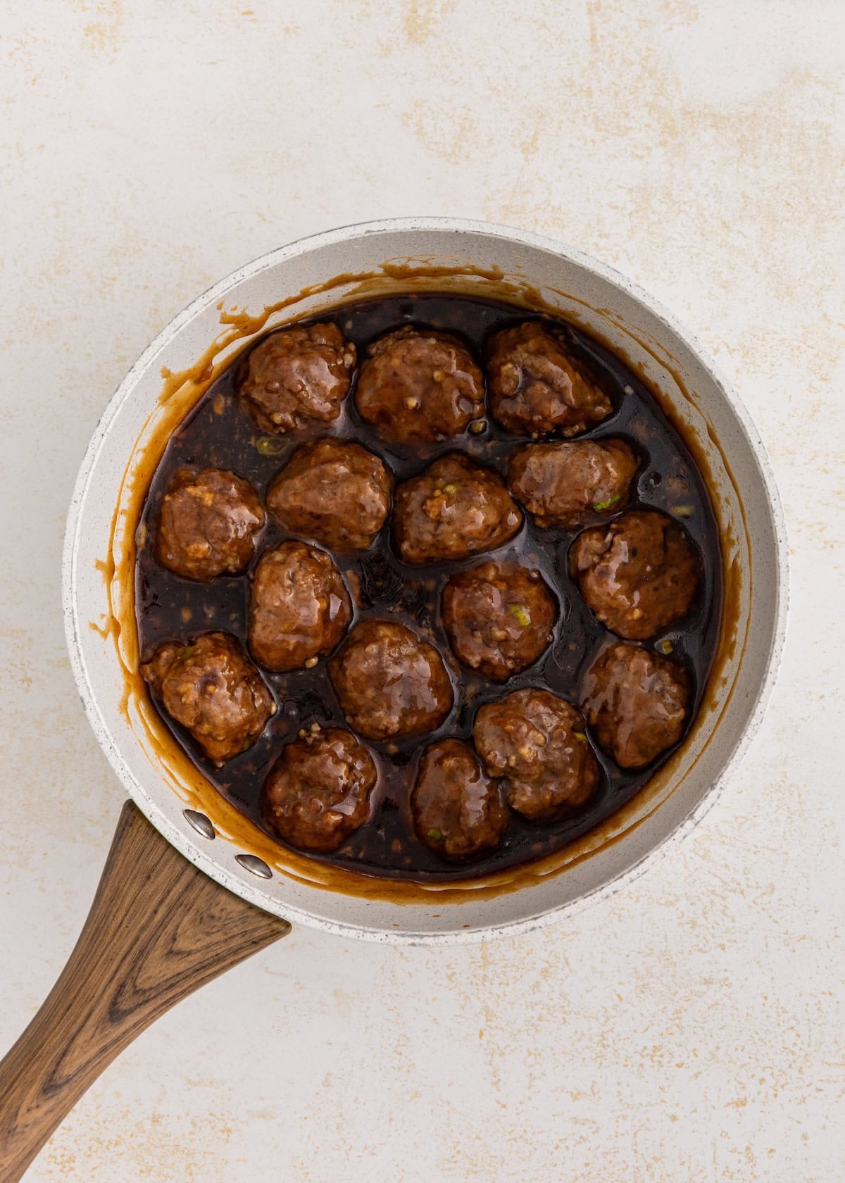 Meatballs in a dark sauce in a white skillet with a wooden handle on a light background.