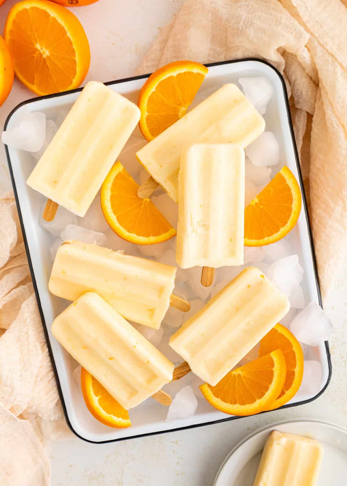 Creamy orange popsicles on a tray with ice cubes and fresh orange slices.