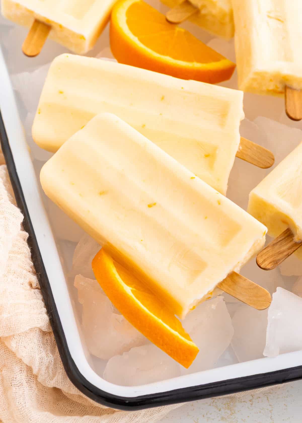 Creamy orange popsicles with orange slices on ice in a white tray.