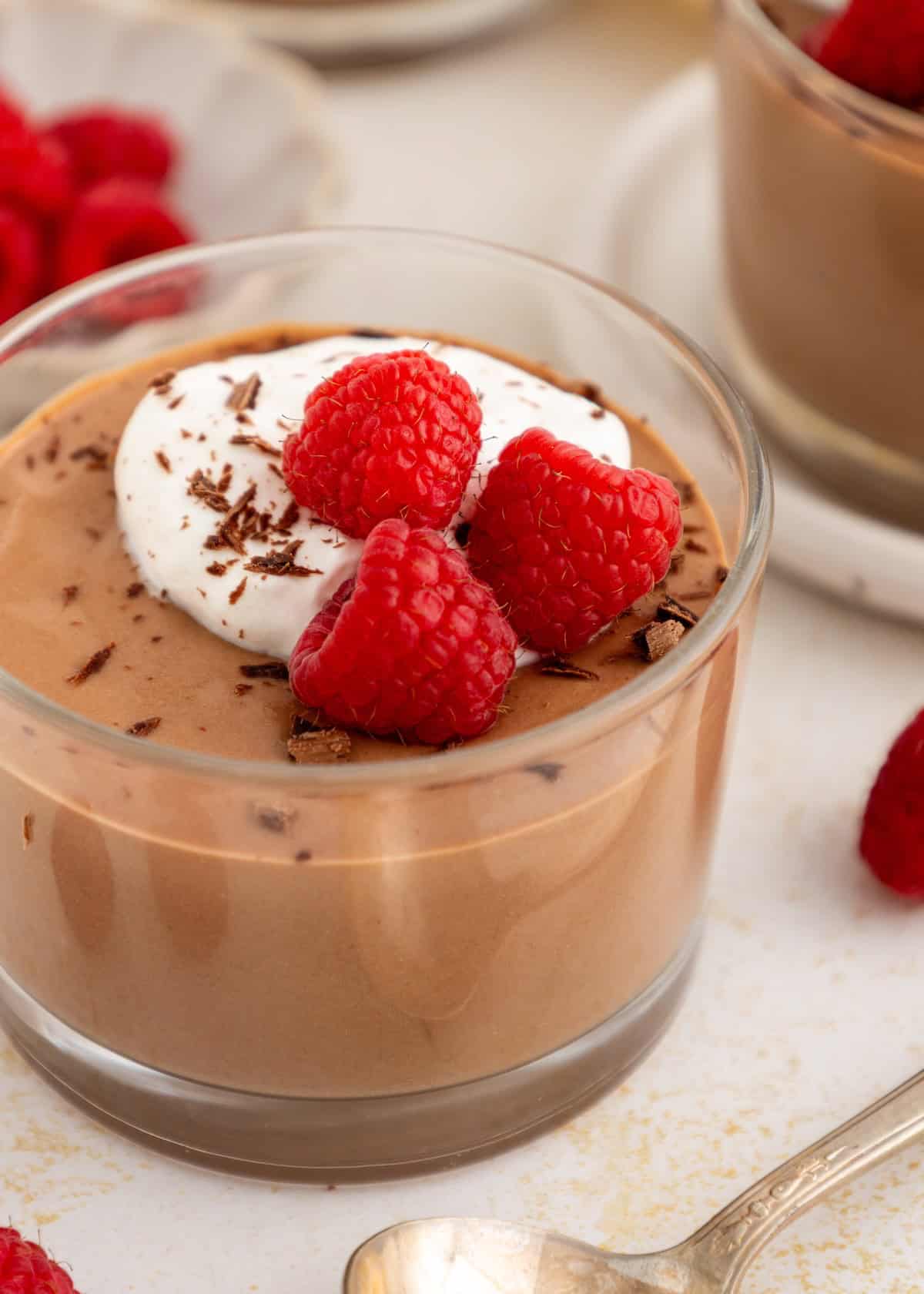 Chocolate mousse in a glass, topped with whipped cream, raspberries, and chocolate shavings.