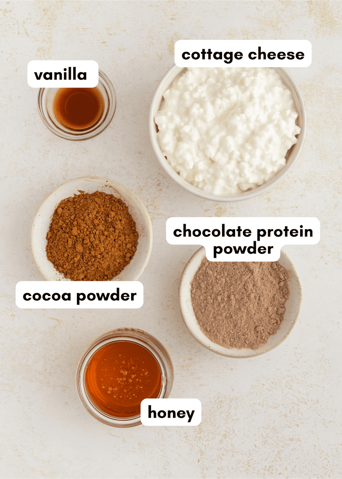 Bowls of cottage cheese, cocoa powder, chocolate protein powder, honey, and vanilla.