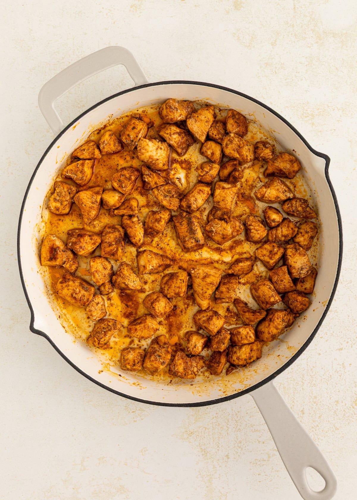 Cooked, seasoned chicken pieces in a white skillet on a light-colored surface.