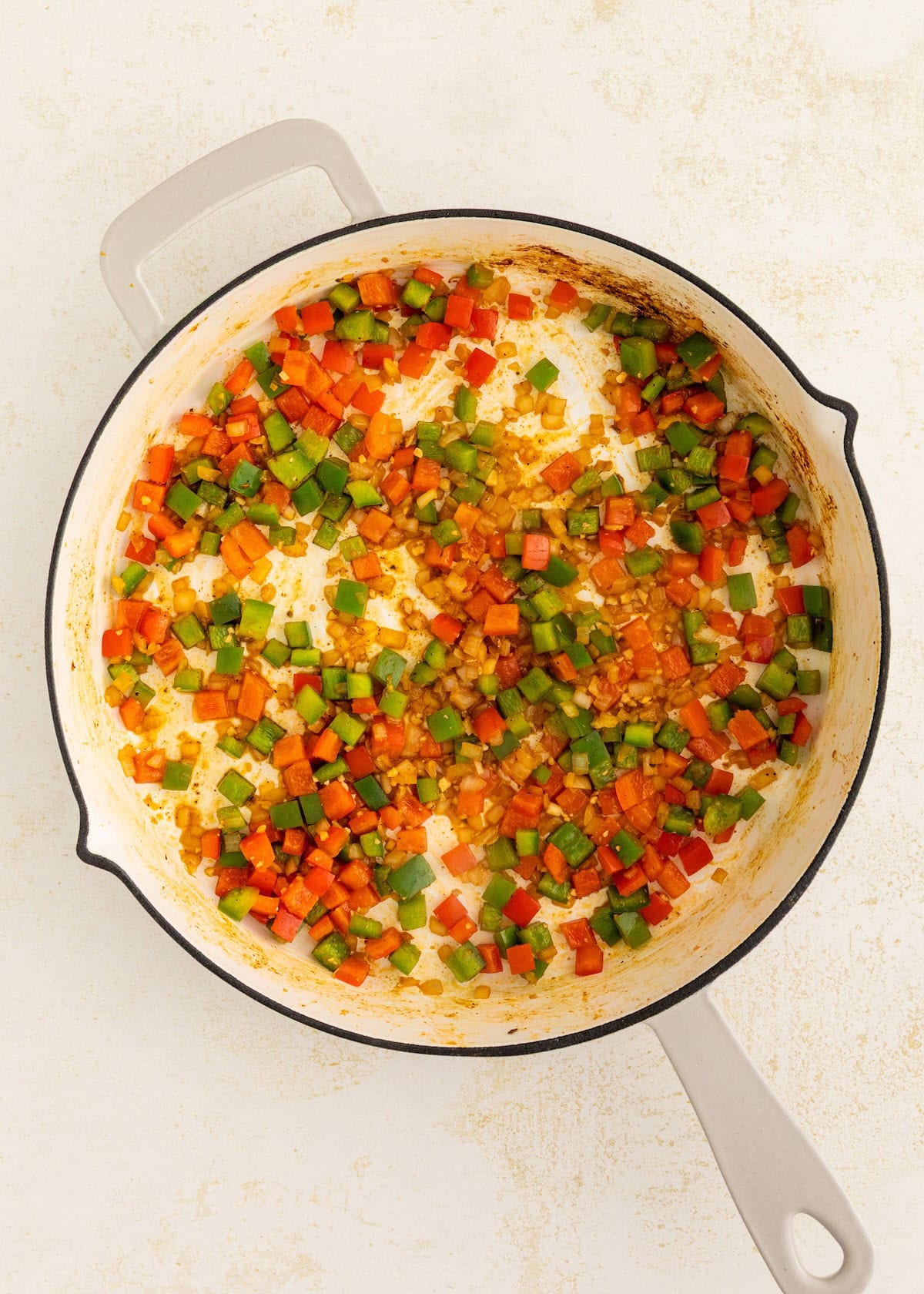 Diced red and green bell peppers and onions sautéing in a white skillet.