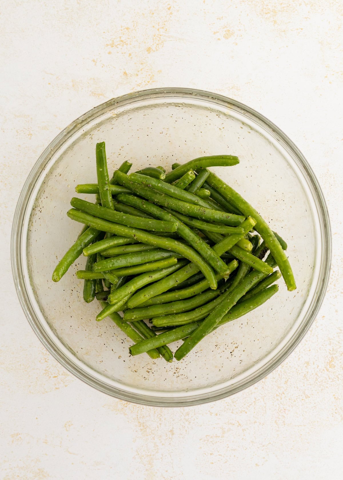Fresh green beans seasoned with pepper in a clear glass bowl on a light surface.