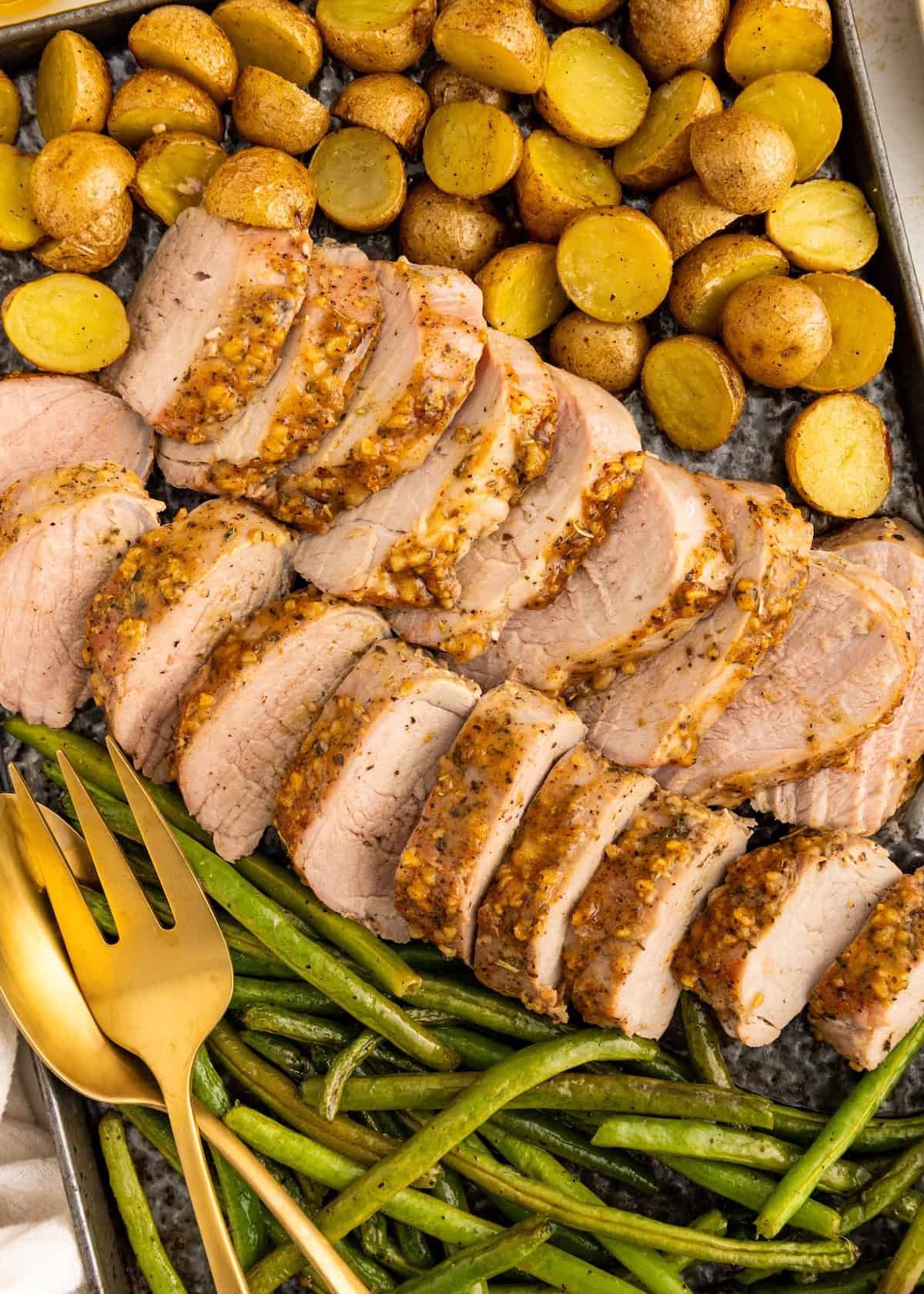 Sliced pork tenderloin, roasted potatoes, and green beans on a tray with serving utensils.