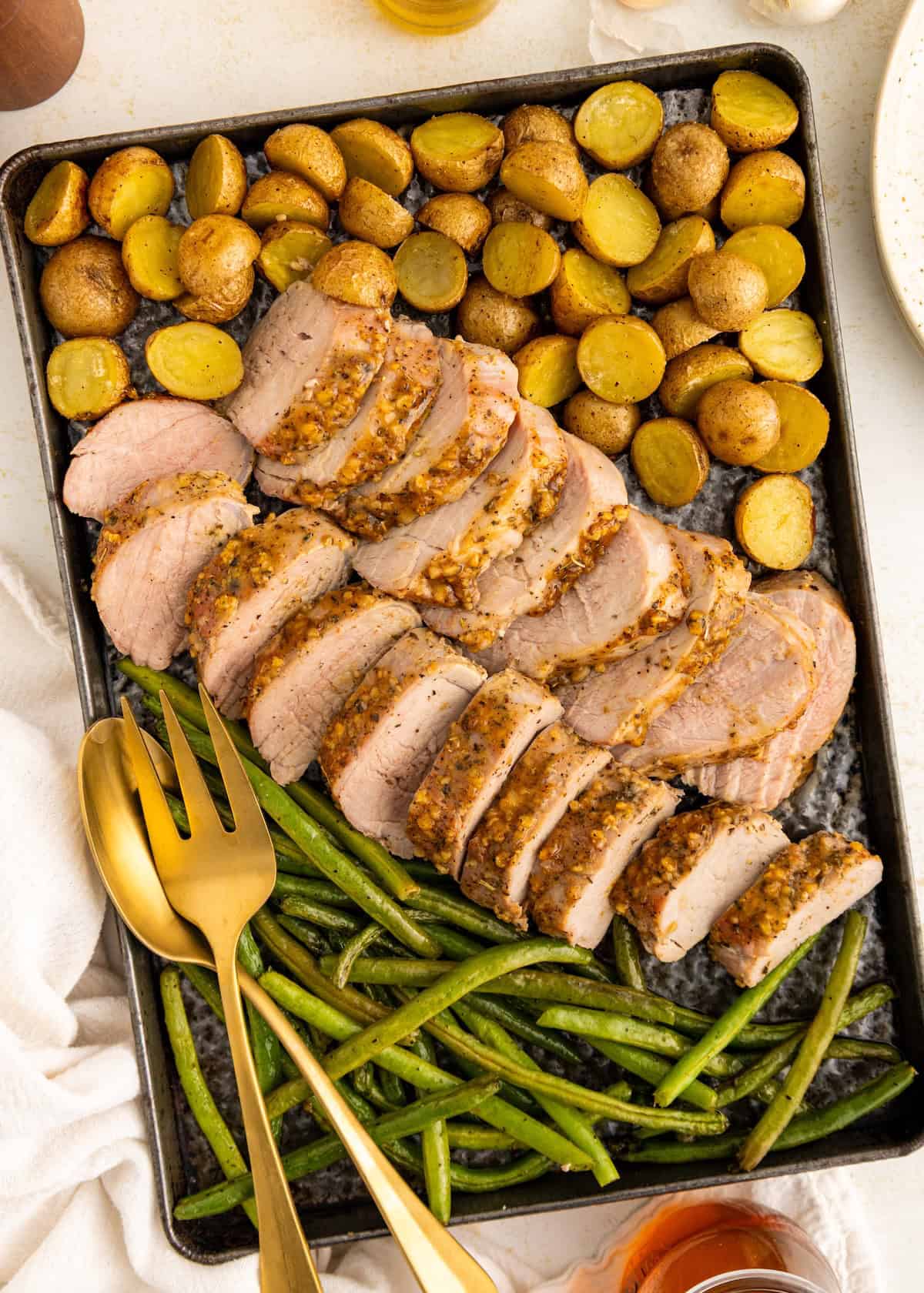 Sliced pork tenderloin, green beans, and roasted potatoes on a baking sheet with serving utensils.