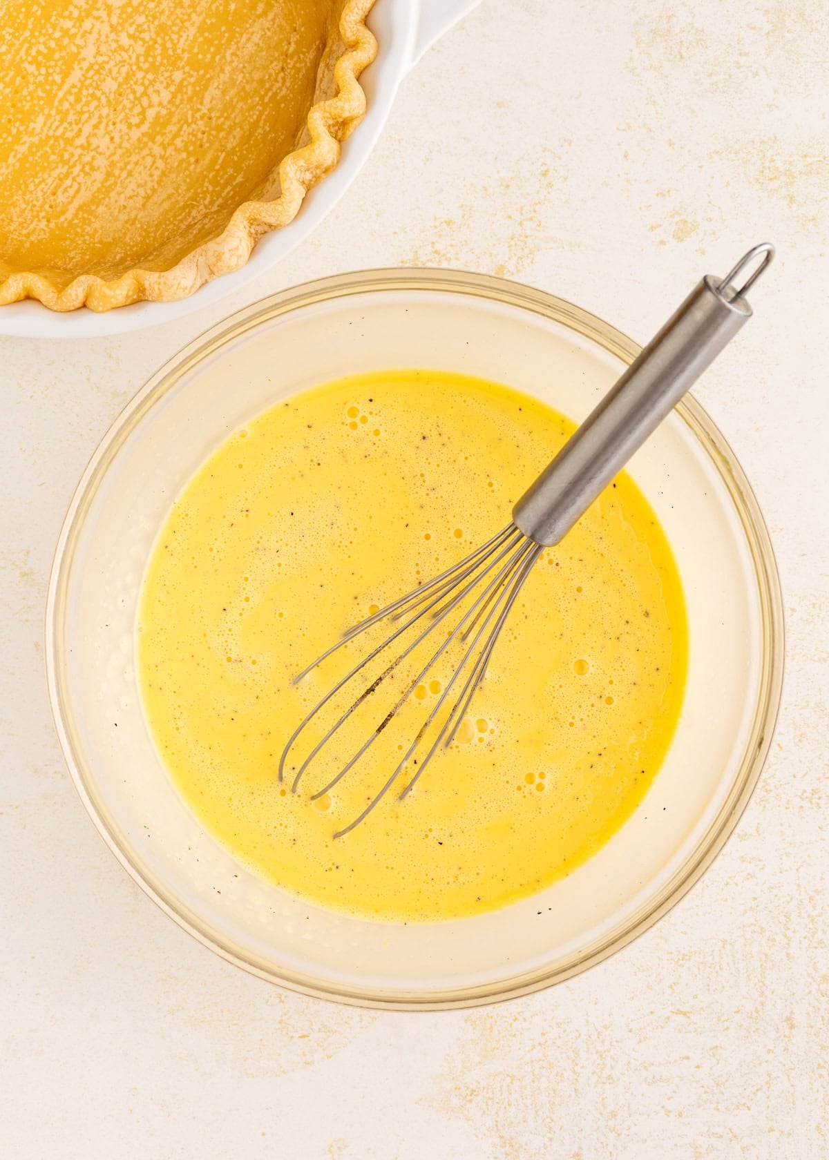 A whisk in a bowl of yellow pie filling next to an unbaked pie crust.