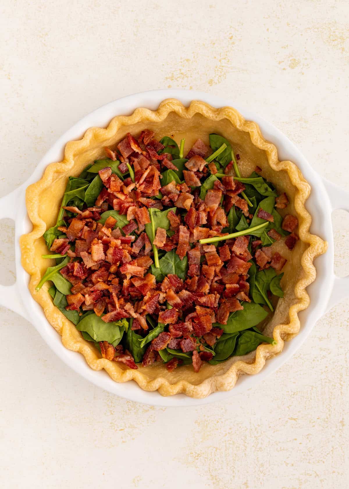 A pie crust filled with fresh spinach and chopped cooked bacon in a white dish.