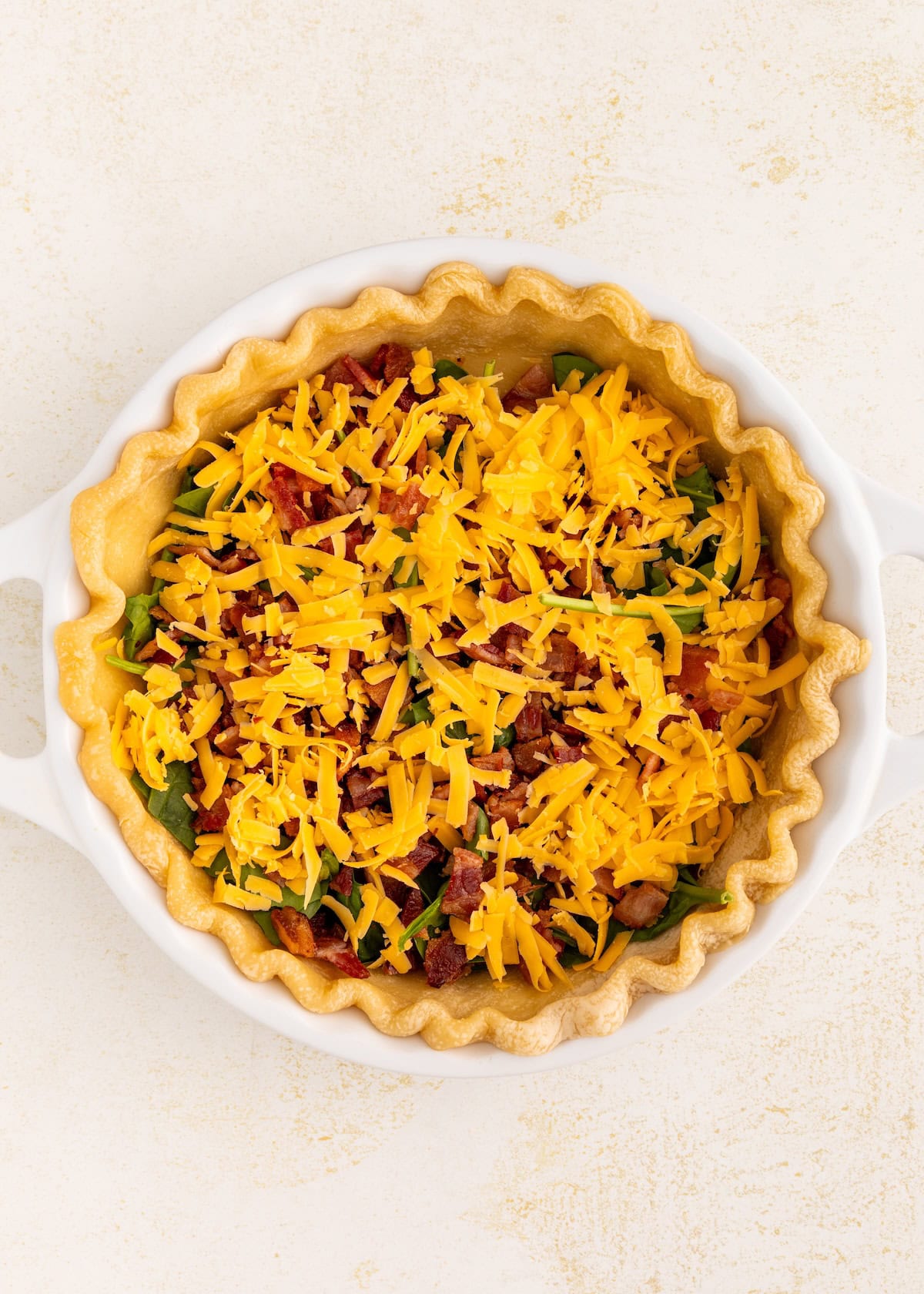 Unbaked pie crust filled with shredded cheese, bacon, and vegetables in a white dish.
