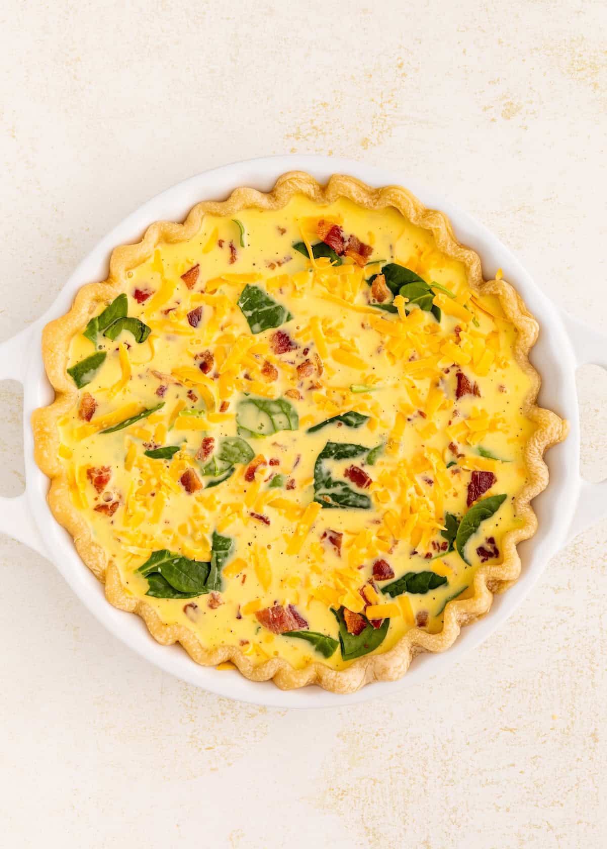Unbaked quiche with spinach, bacon, and cheese in a white pie dish on a light background.
