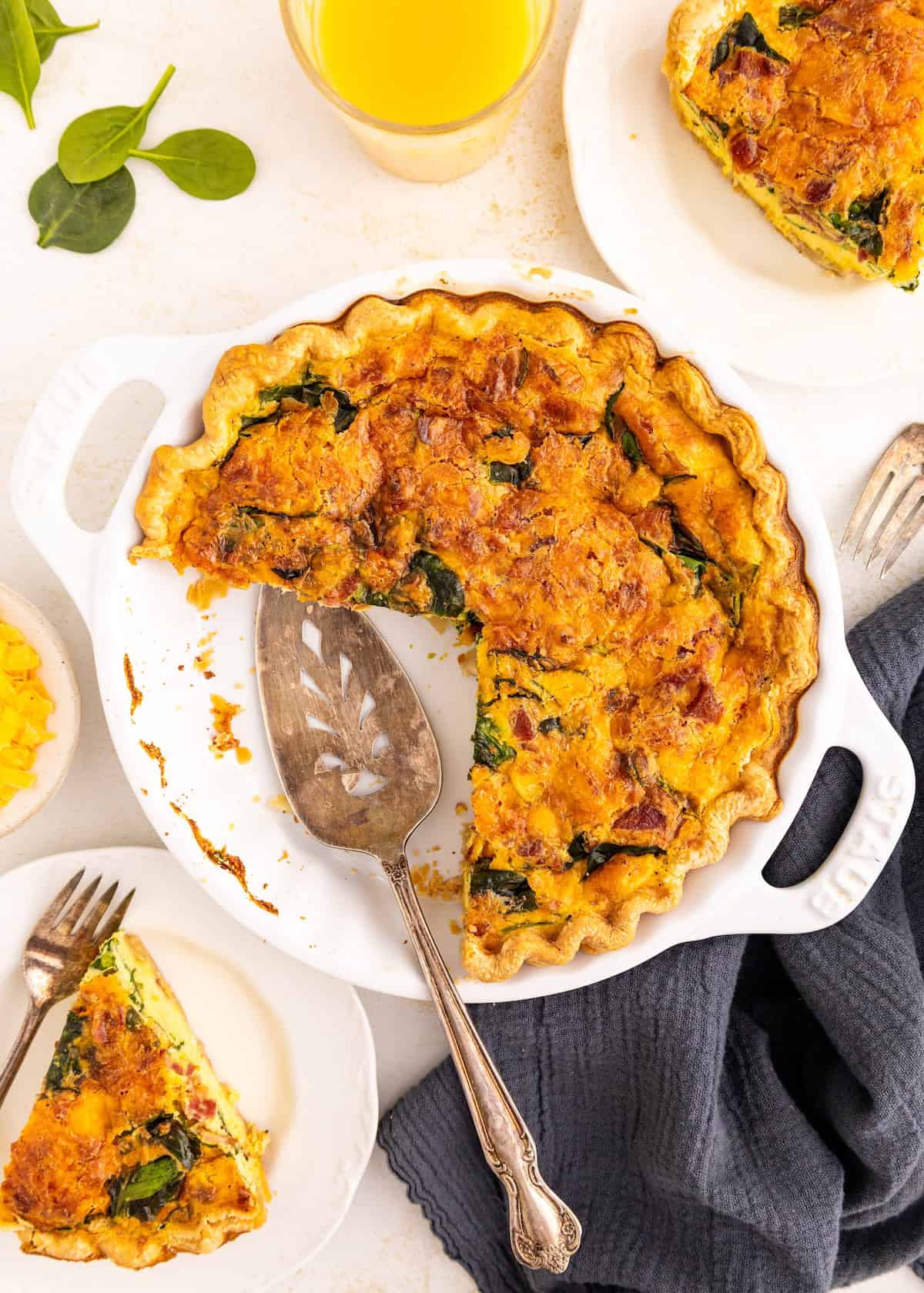 A sliced spinach and cheese quiche with serving utensils and plates on a white table.