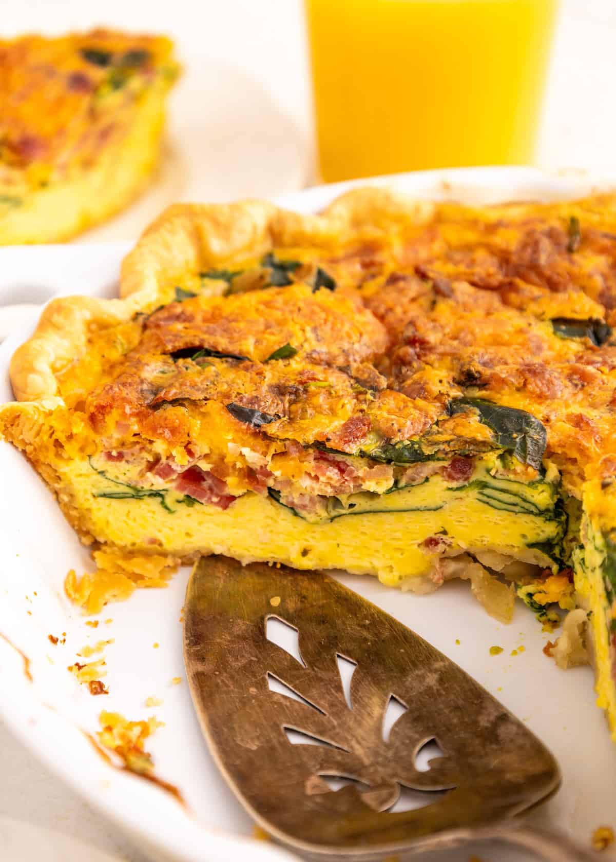 A slice of spinach and bacon quiche in a white dish with a glass of orange juice.