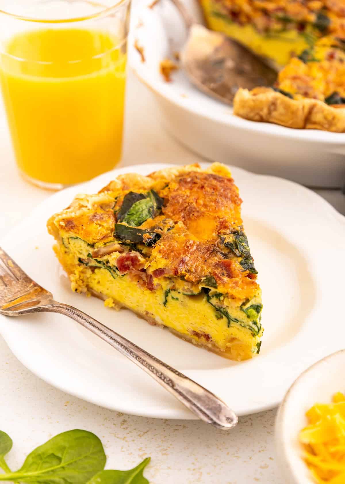 A slice of vegetable quiche on a white plate with a glass of orange juice beside it.