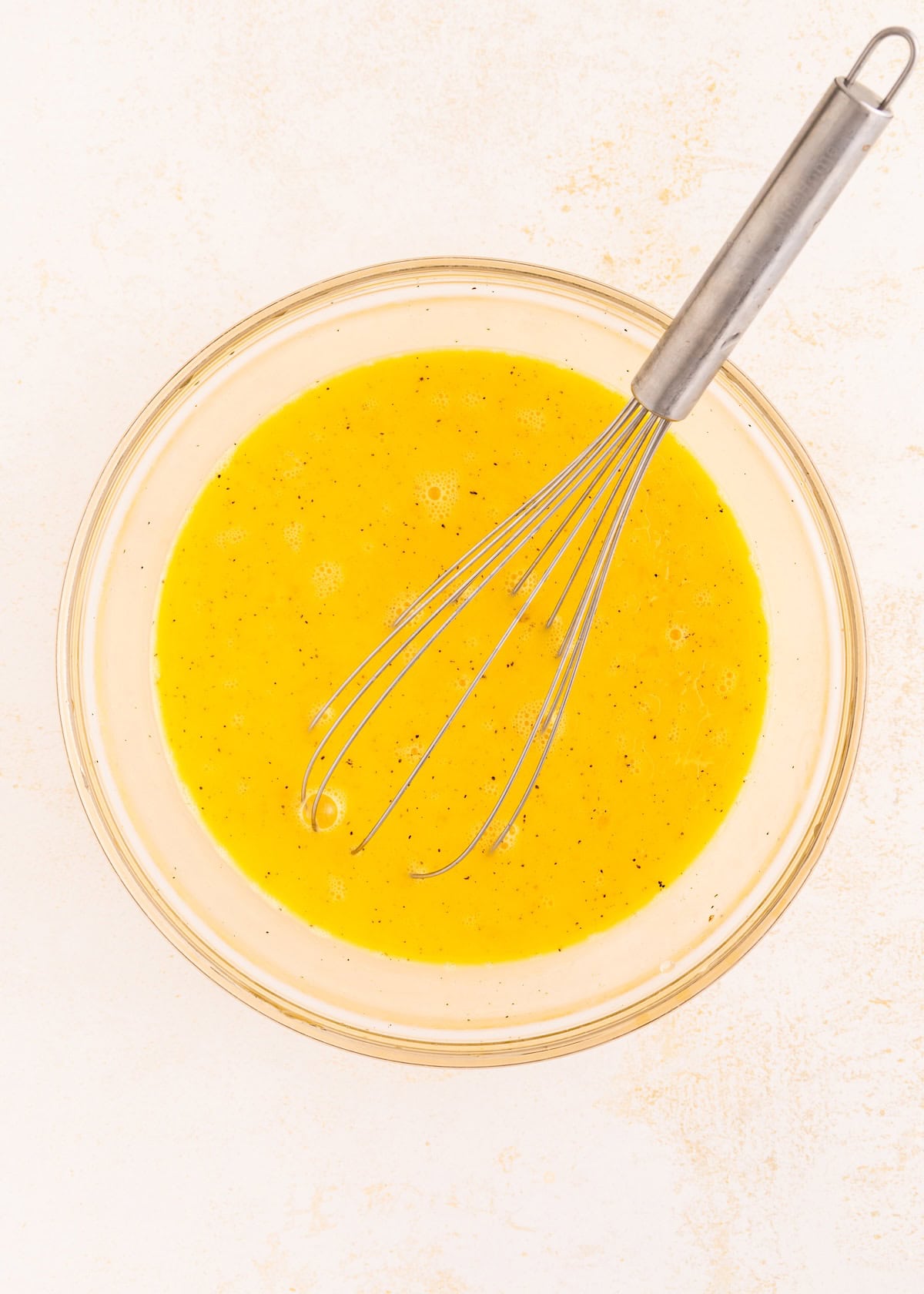 Whisk in a glass bowl with beaten eggs on a light surface.