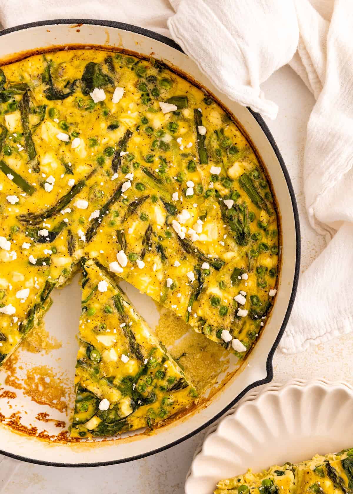 Egg and vegetable frittata in a skillet with a few slices removed, topped with crumbled cheese.