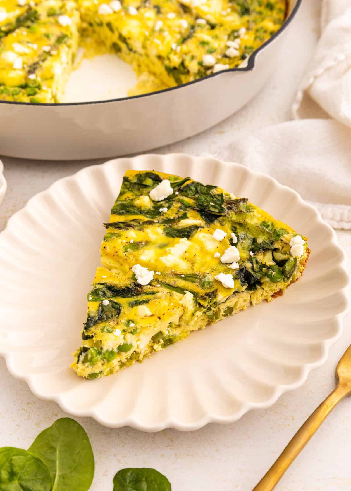 A slice of spinach and feta frittata on a white plate with a gold fork beside it.
