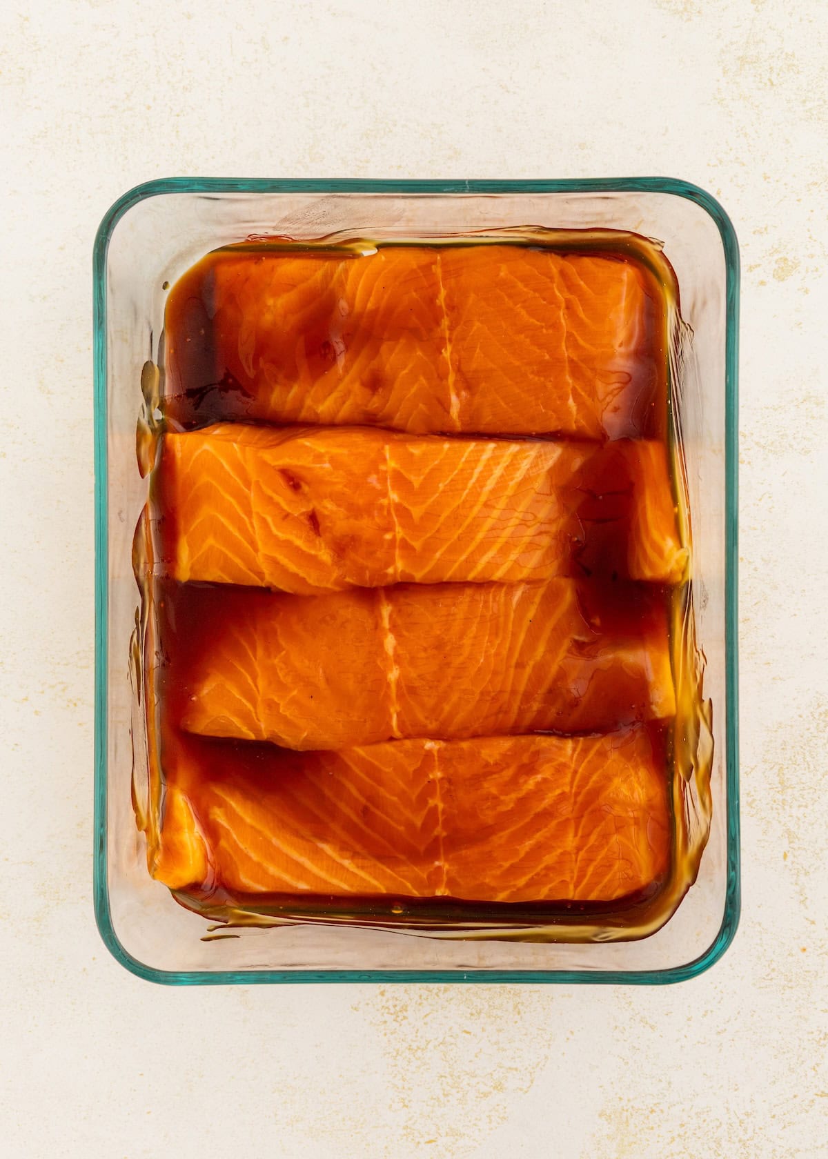 Four raw salmon fillets marinating in a glass dish with a dark liquid marinade.