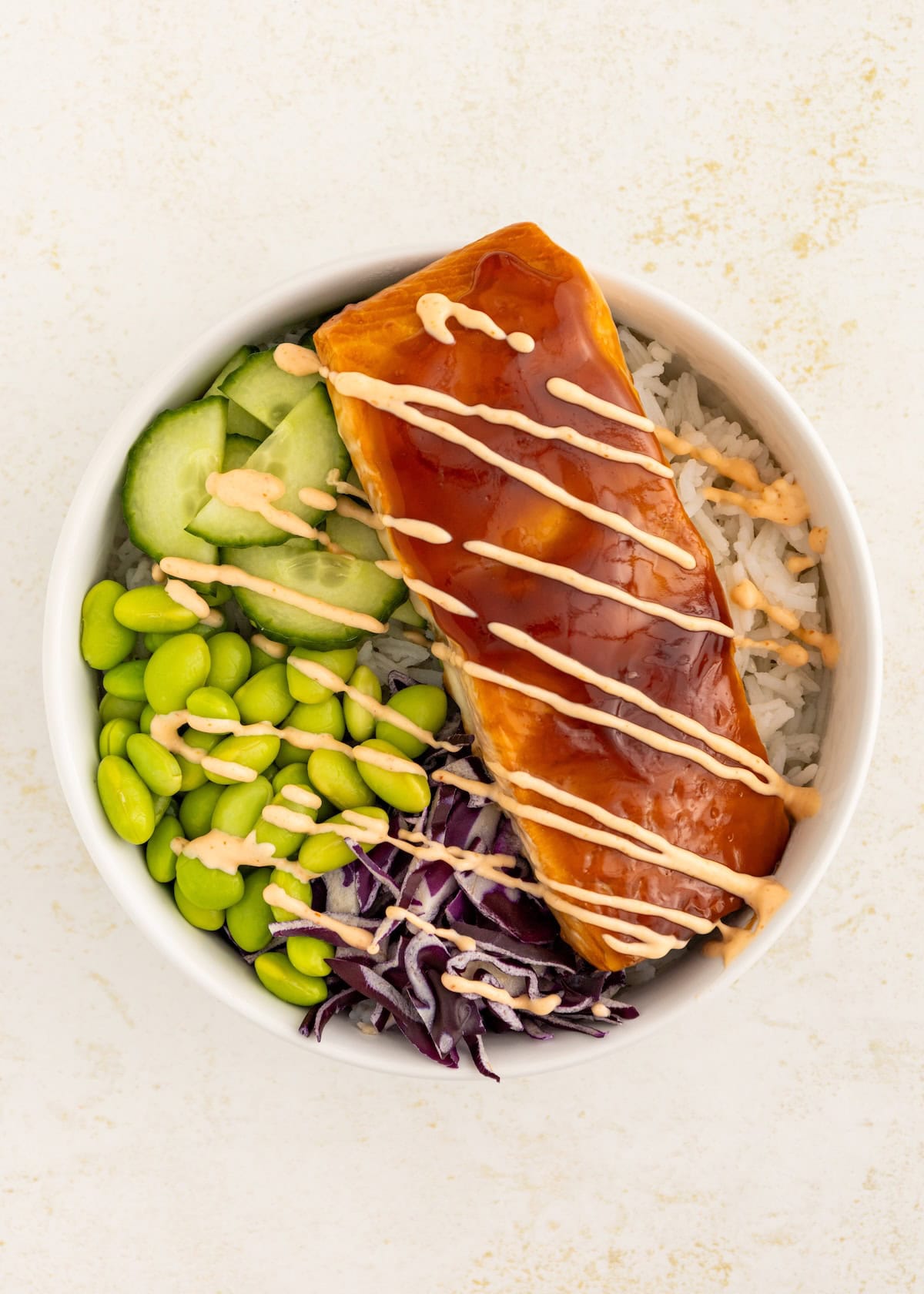 A bowl with glazed salmon, rice, edamame, cucumber, cabbage, and a drizzle of sauce.