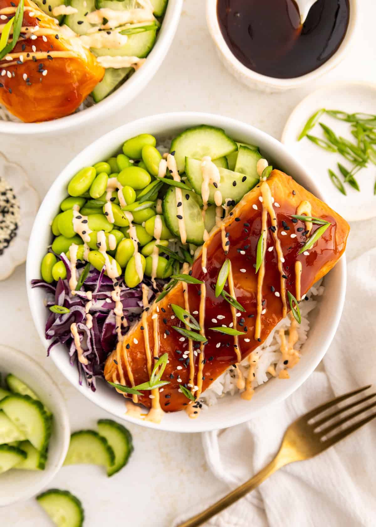 Bowl with glazed salmon, rice, edamame, cucumber, cabbage, and sauce drizzles on top.
