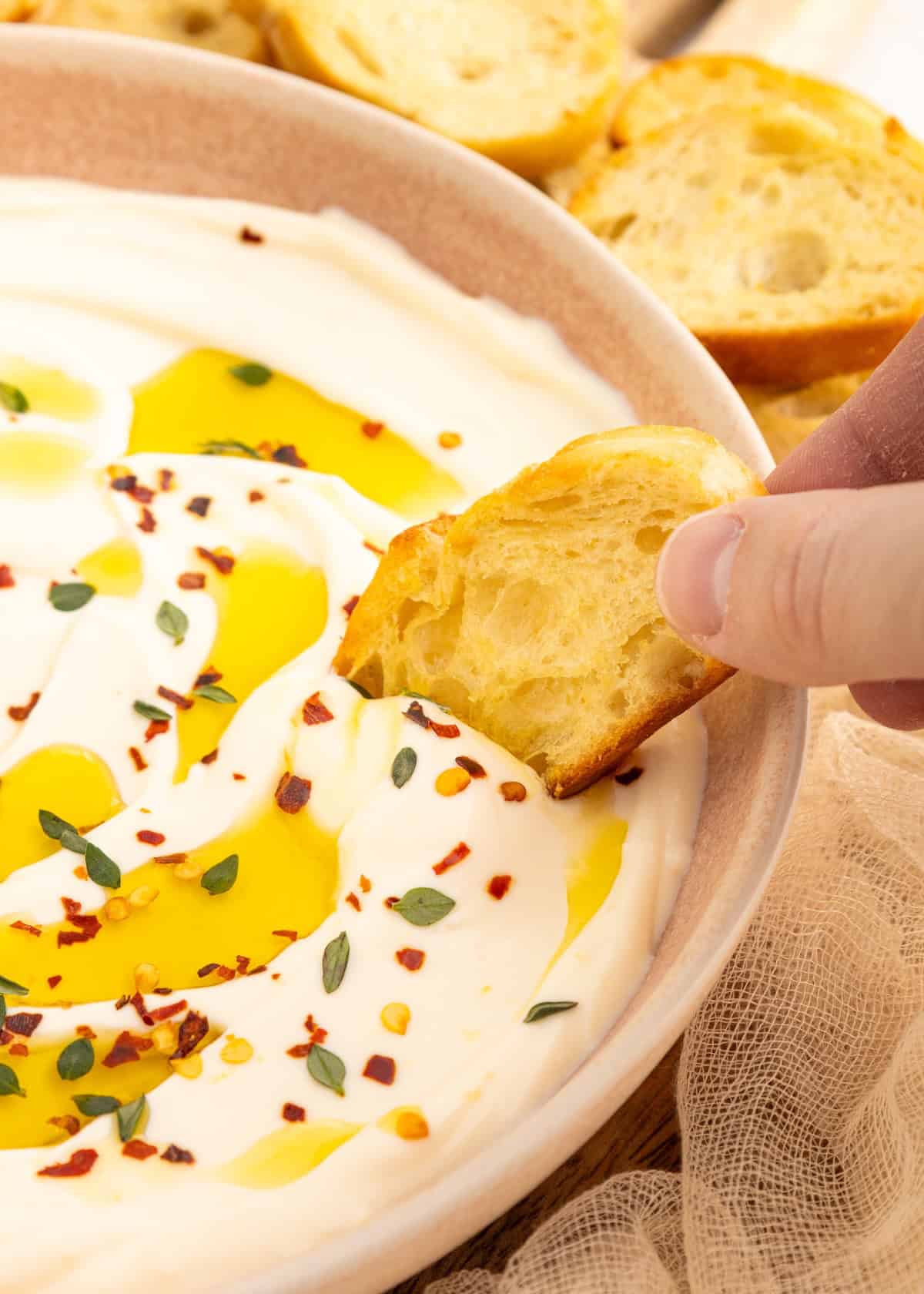 Hand dipping bread into creamy dip topped with olive oil, herbs, and chili flakes.