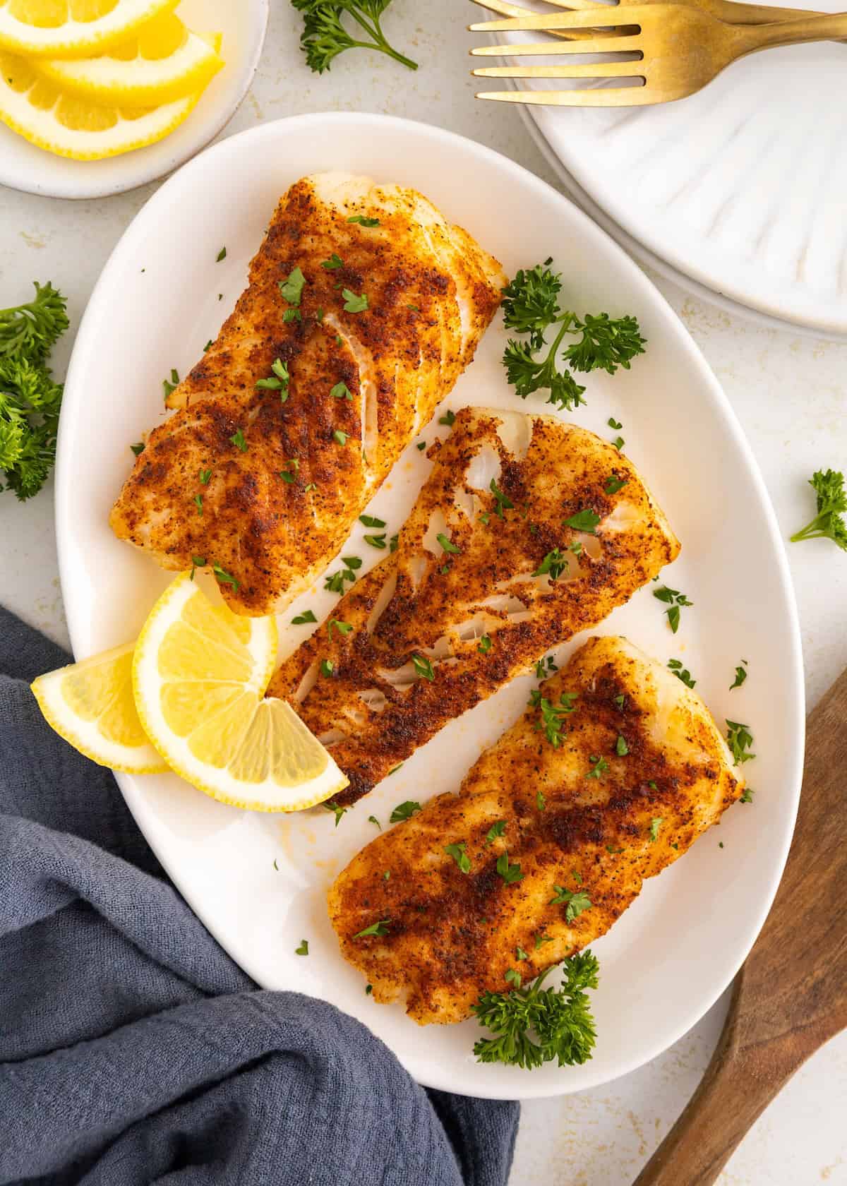 Three seasoned air fryer cod fillets with lemon slices and parsley on a white oval plate.