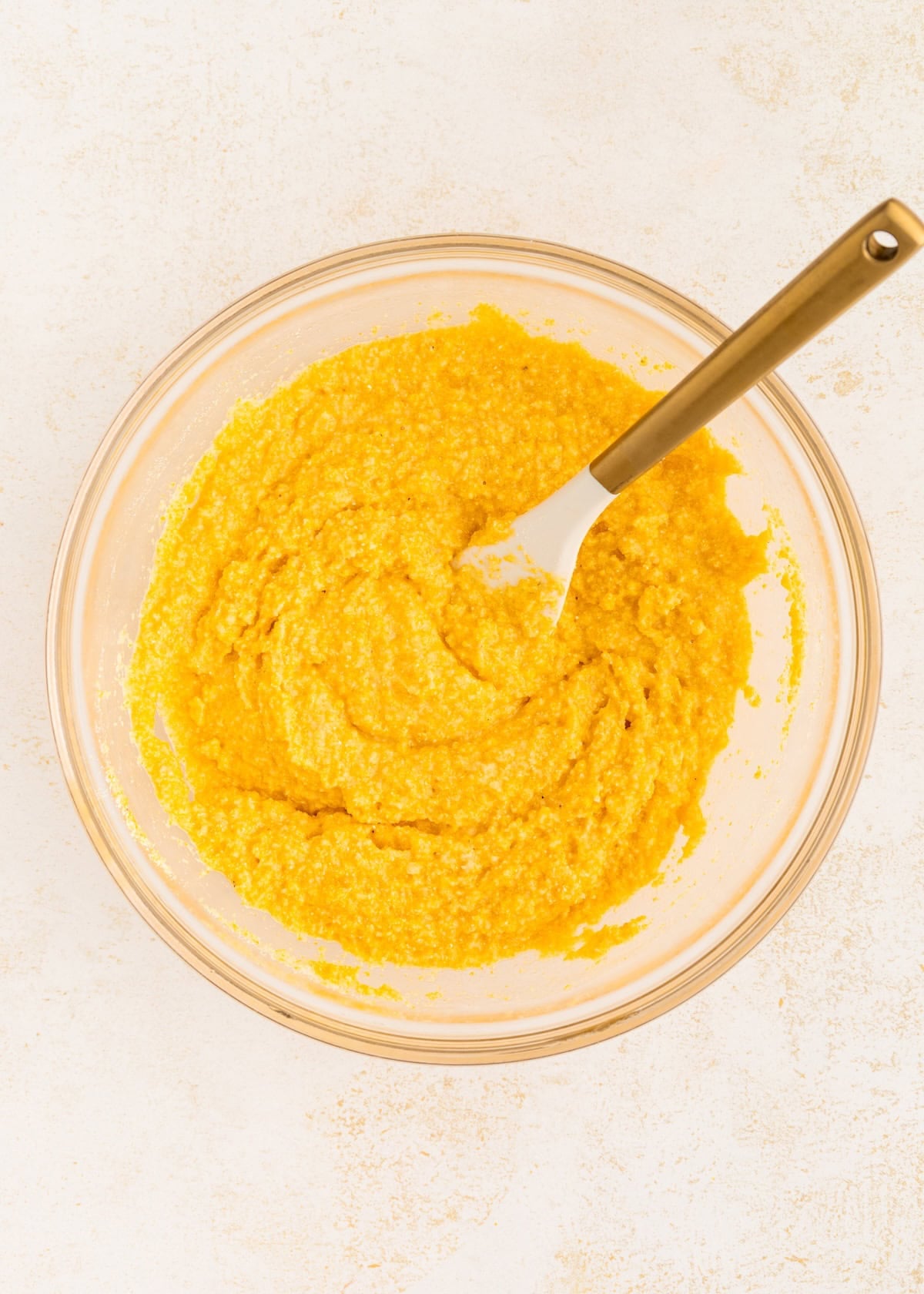 A glass bowl with yellow batter and a spatula on a light-colored surface.
